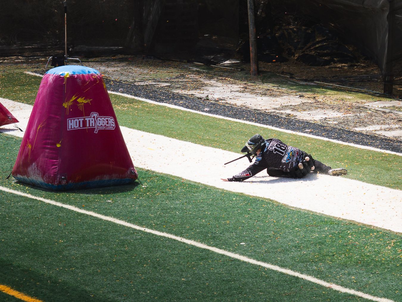 Player crawling on turf field during paintball game near inflatable bunker obstacle.