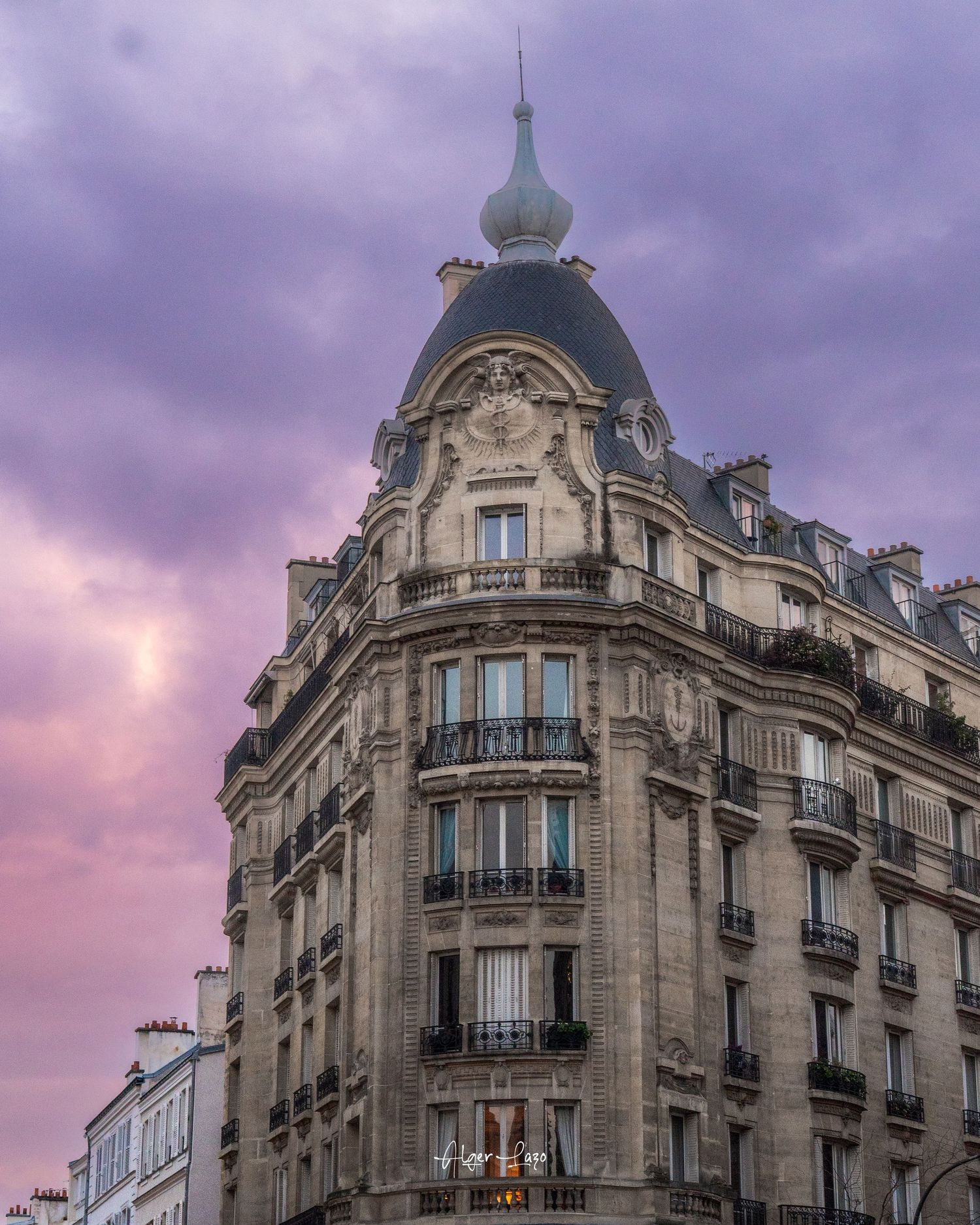 Ornate Haussmann-style Parisian building corner with iron balconies and domed turret under dramatic purple sunset sky.