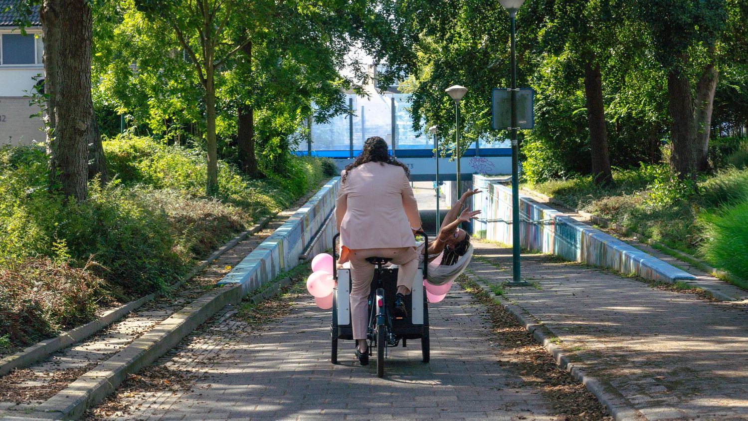 Cyclist rides cargo bike on tree-lined path with blue building in background on sunny day.