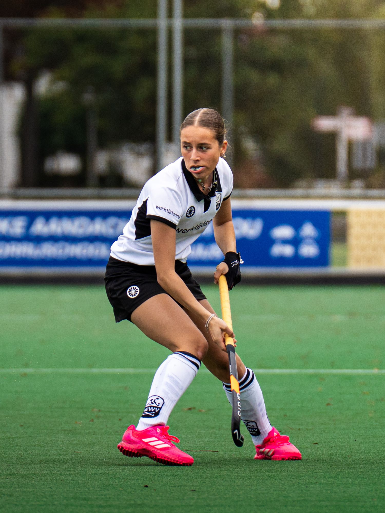 Field hockey player in white uniform crouches low to control ball with stick during outdoor match.
