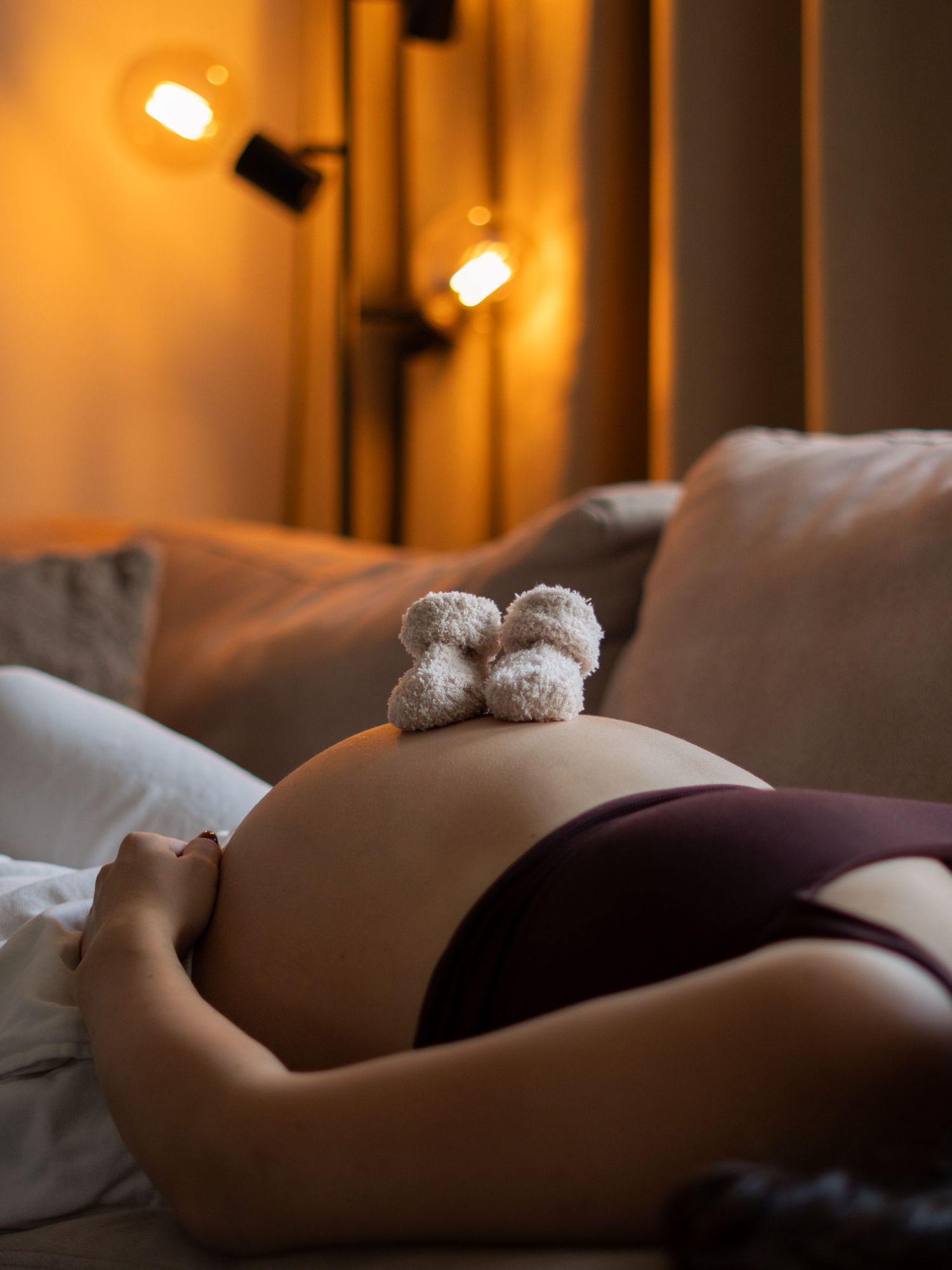 A pair of tiny knitted booties rests on a pregnant belly in warm lighting with cozy bedding.
