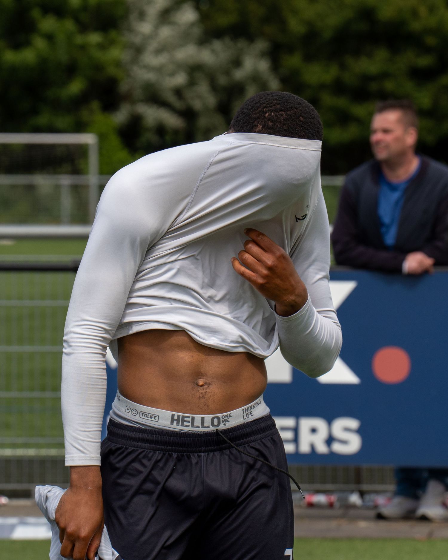 A person in a white long-sleeve shirt and dark pants lifts their shirt to show their stomach during an outdoor event.
