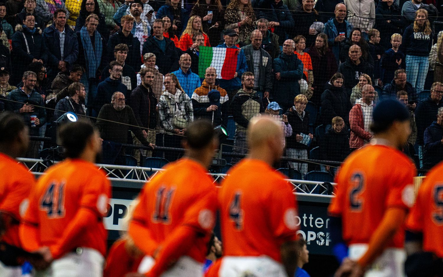 Soccer fans celebrate in a crowded stadium with orange-clad players visible on the field during a match.