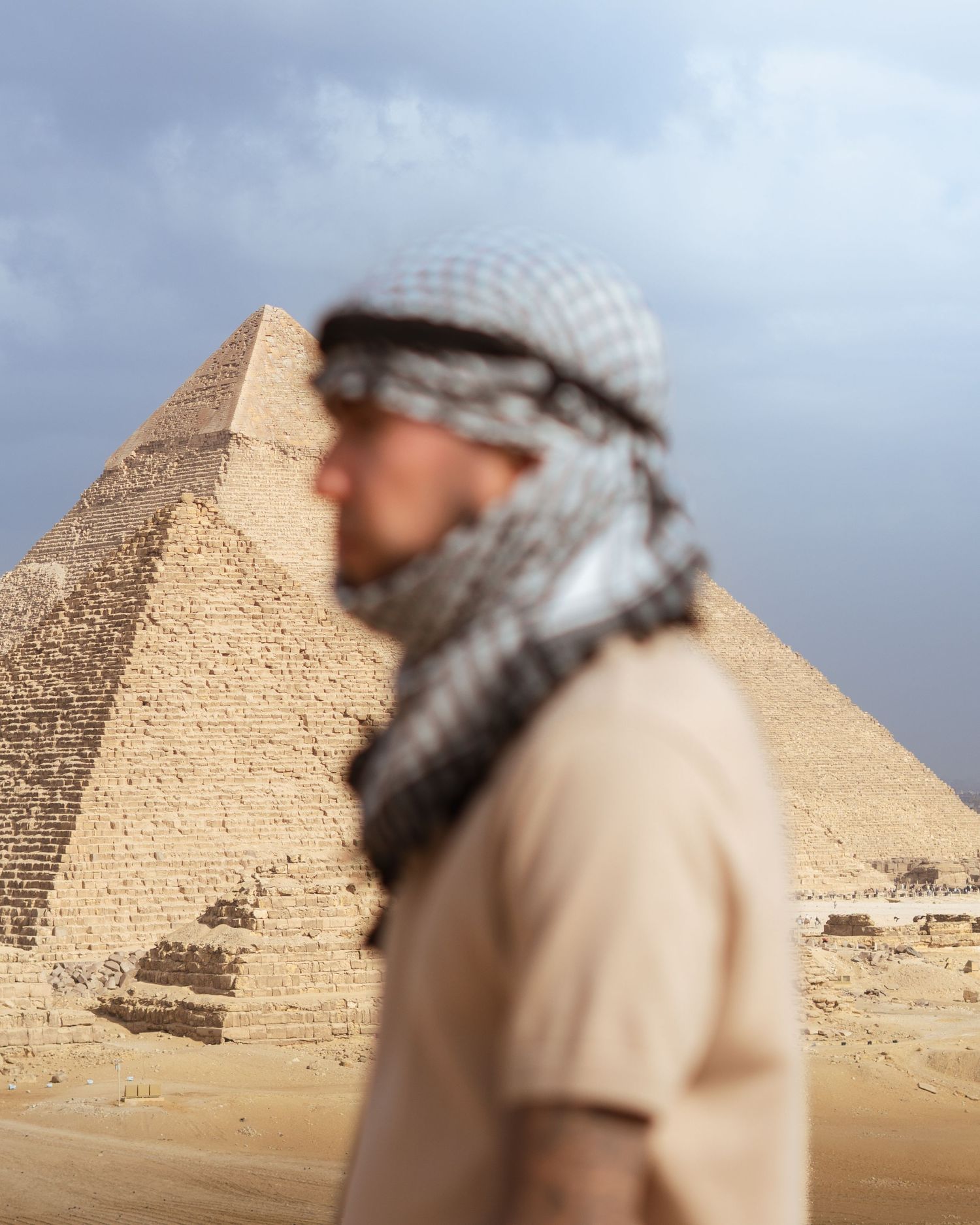 A person wearing traditional Middle Eastern headwear stands in front of the ancient pyramids of Giza in Egypt.