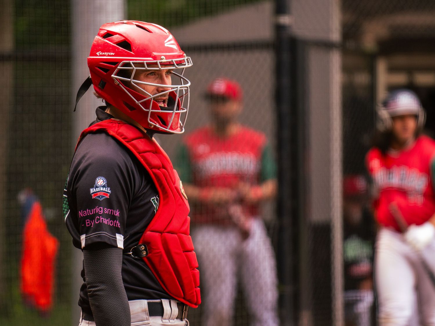 Baseball catcher in red protective gear crouches behind home plate during a game.