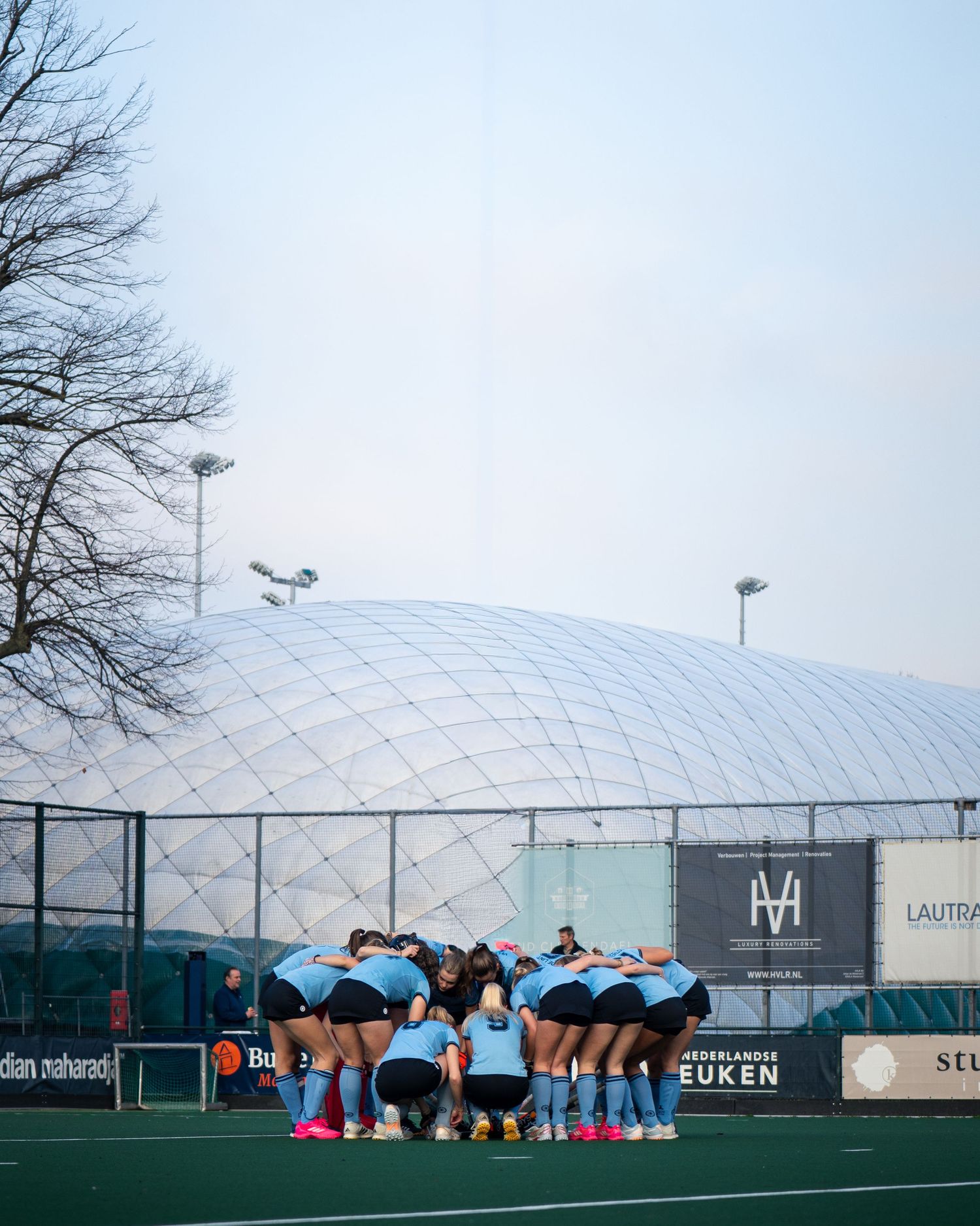 Athletes stretch and warm up on outdoor sports field next to large white dome structure during winter training session.