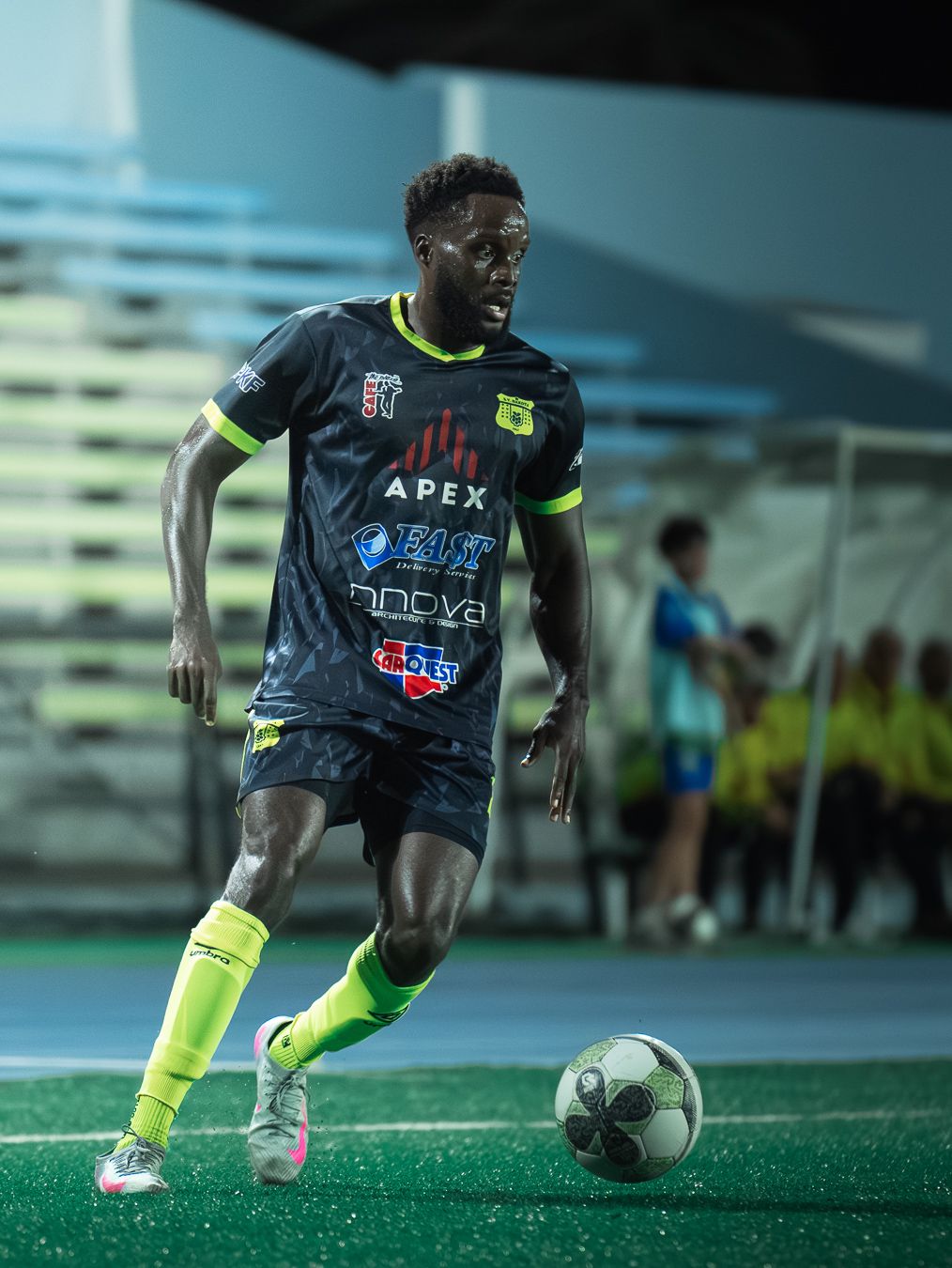 Soccer player in dark APEX jersey dribbles ball on field under night lights wearing neon green socks and cleats.
