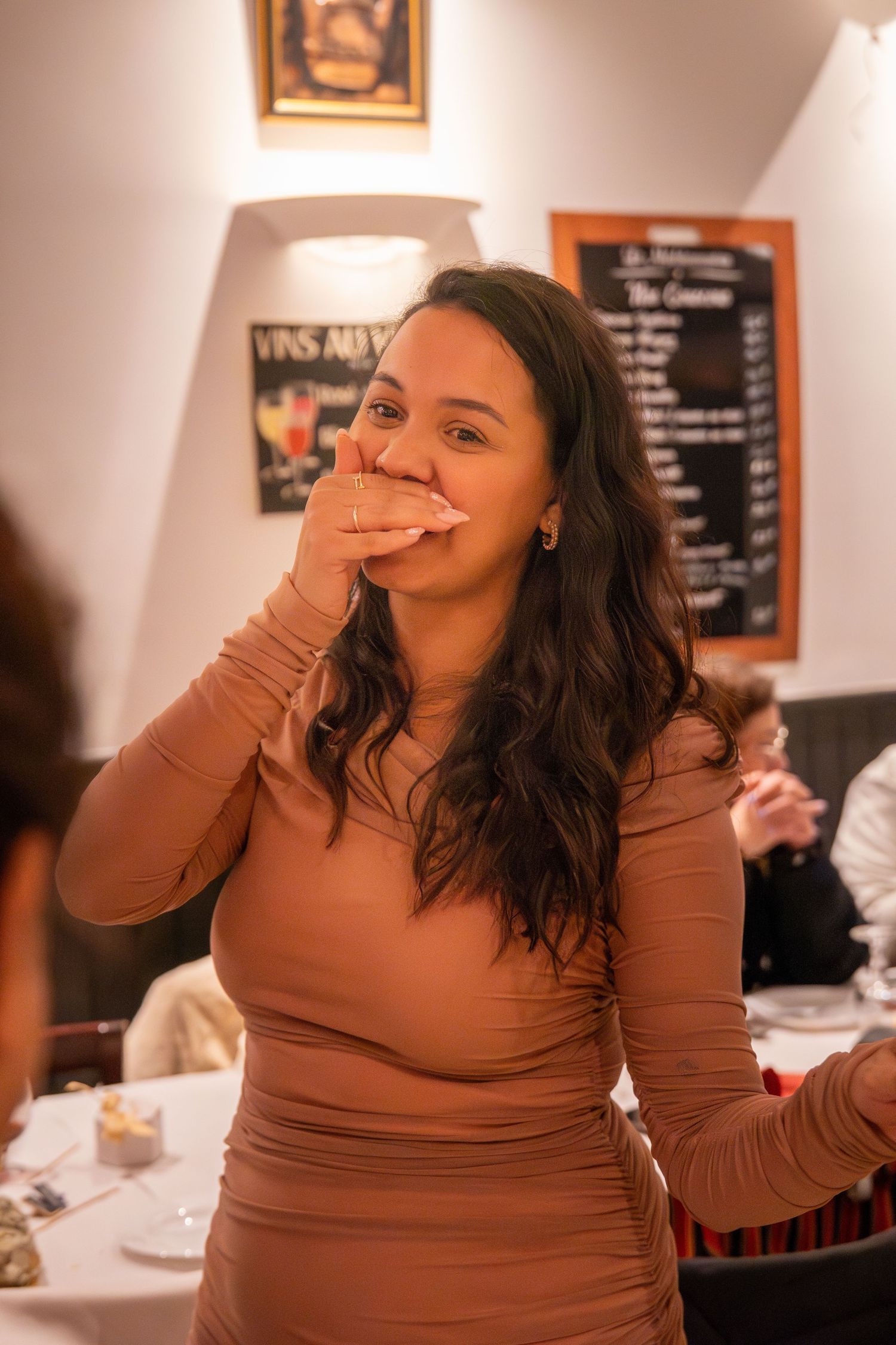 A person in a brown fitted dress laughs while covering their mouth at an indoor gathering.