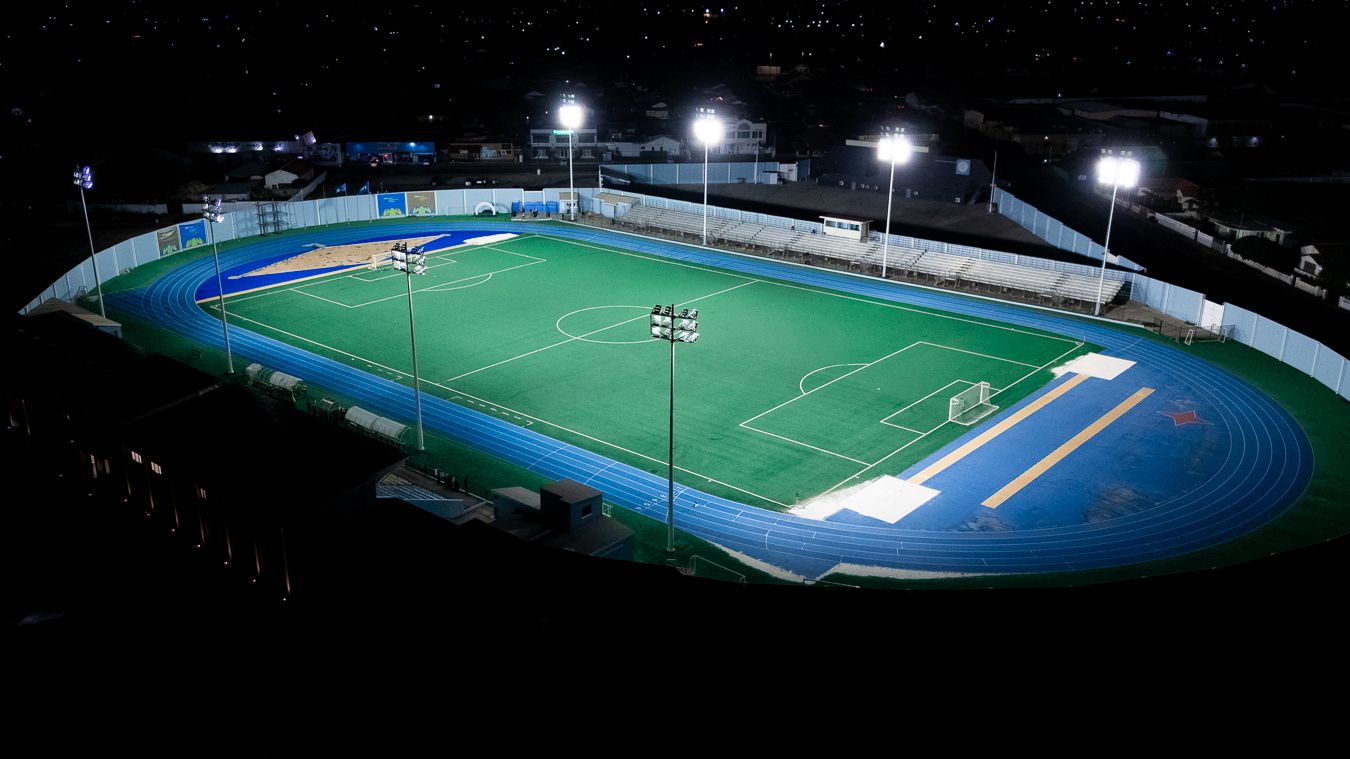 Aerial night view of an illuminated soccer field with blue running track under bright stadium lights.