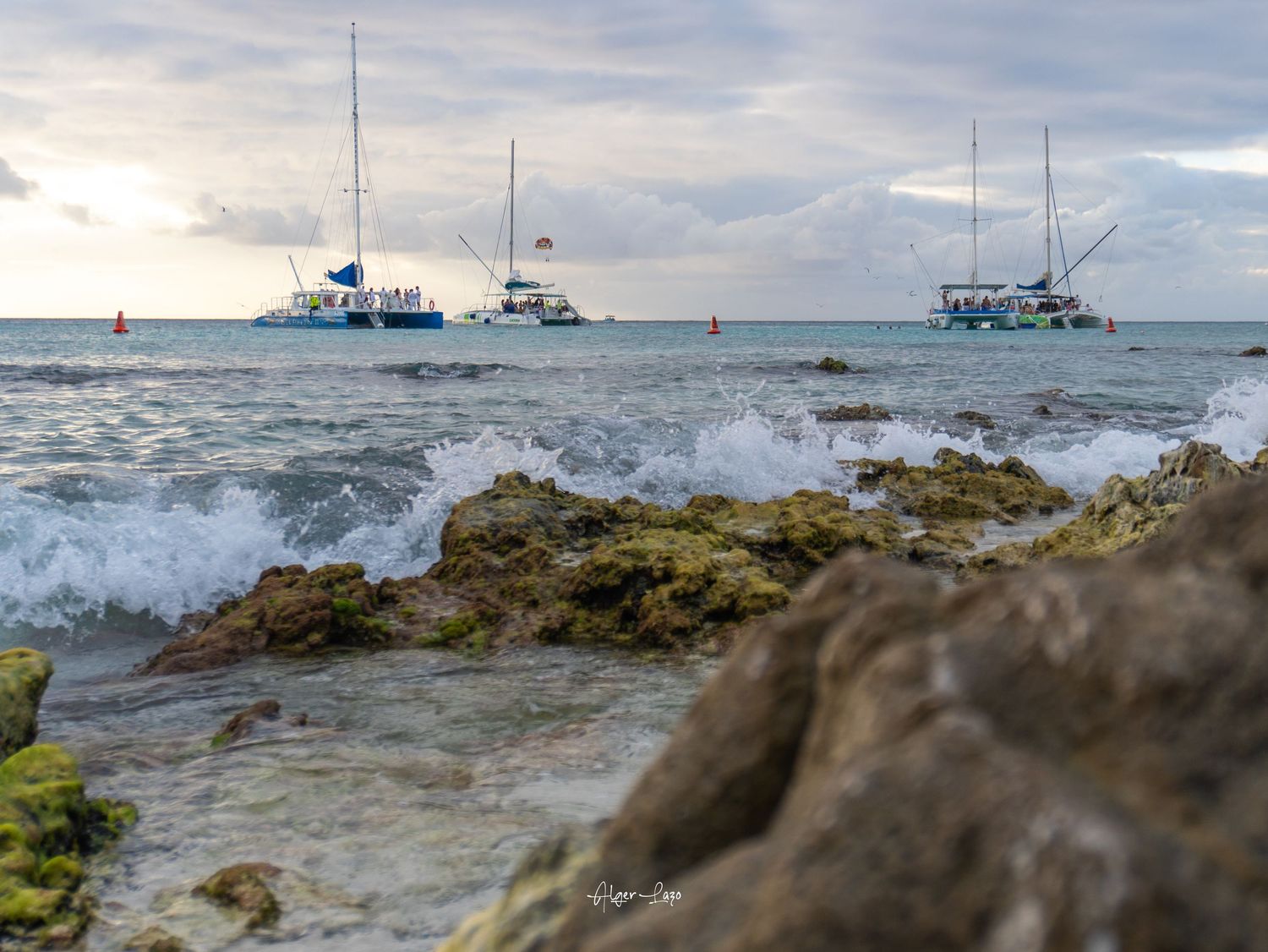 Rocky shoreline with waves crashing, three sailboats anchored in calm Caribbean waters under a cloudy sunset sky.