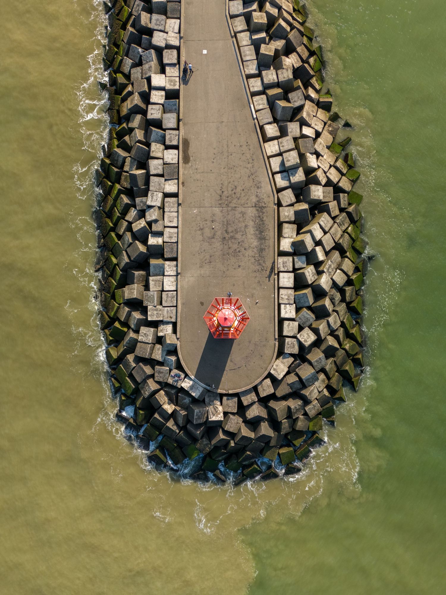 Aerial view of a stone breakwater jetty with tetrapod blocks and a red lighthouse at the tip, surrounded by green water.