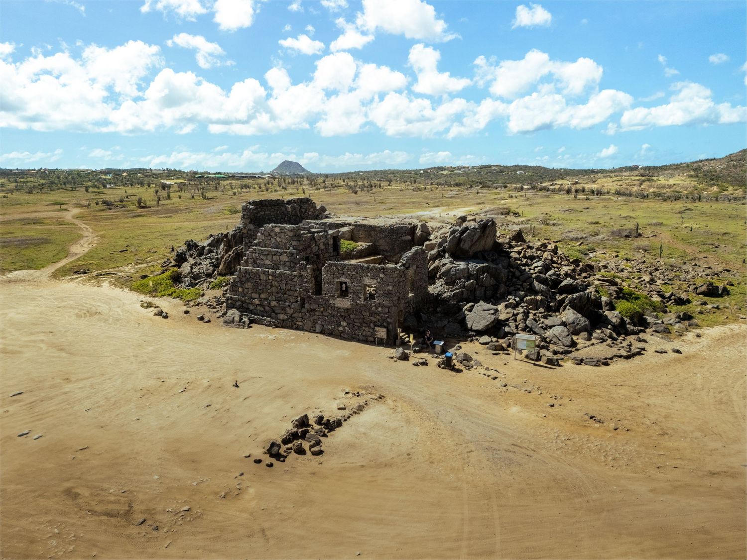 Ancient stone ruins stand in an arid desert landscape under a partly cloudy sky with rocky terrain in the background.