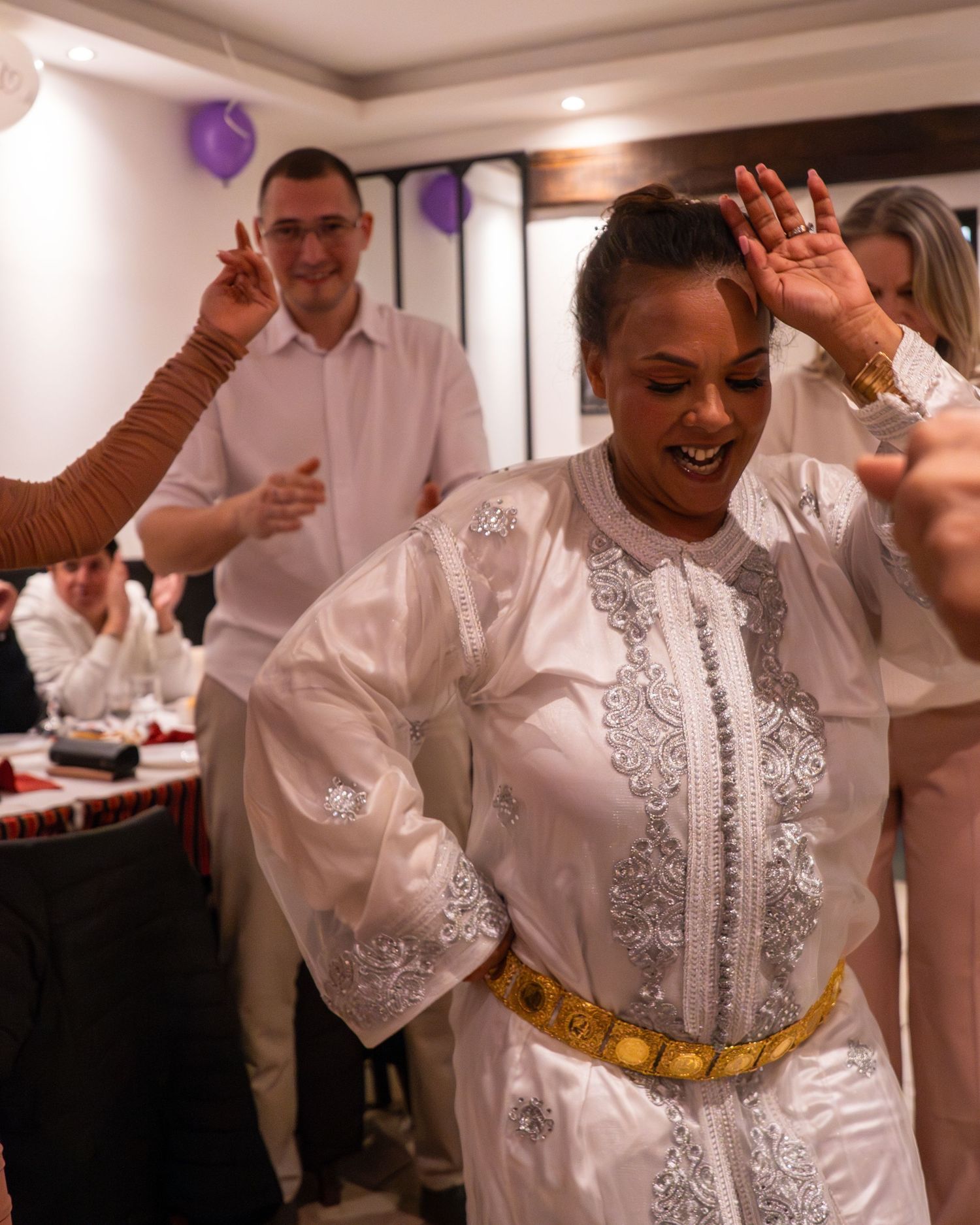 A person in a white embroidered kaftan with gold belt dances joyfully at a celebration with purple balloons in the background.