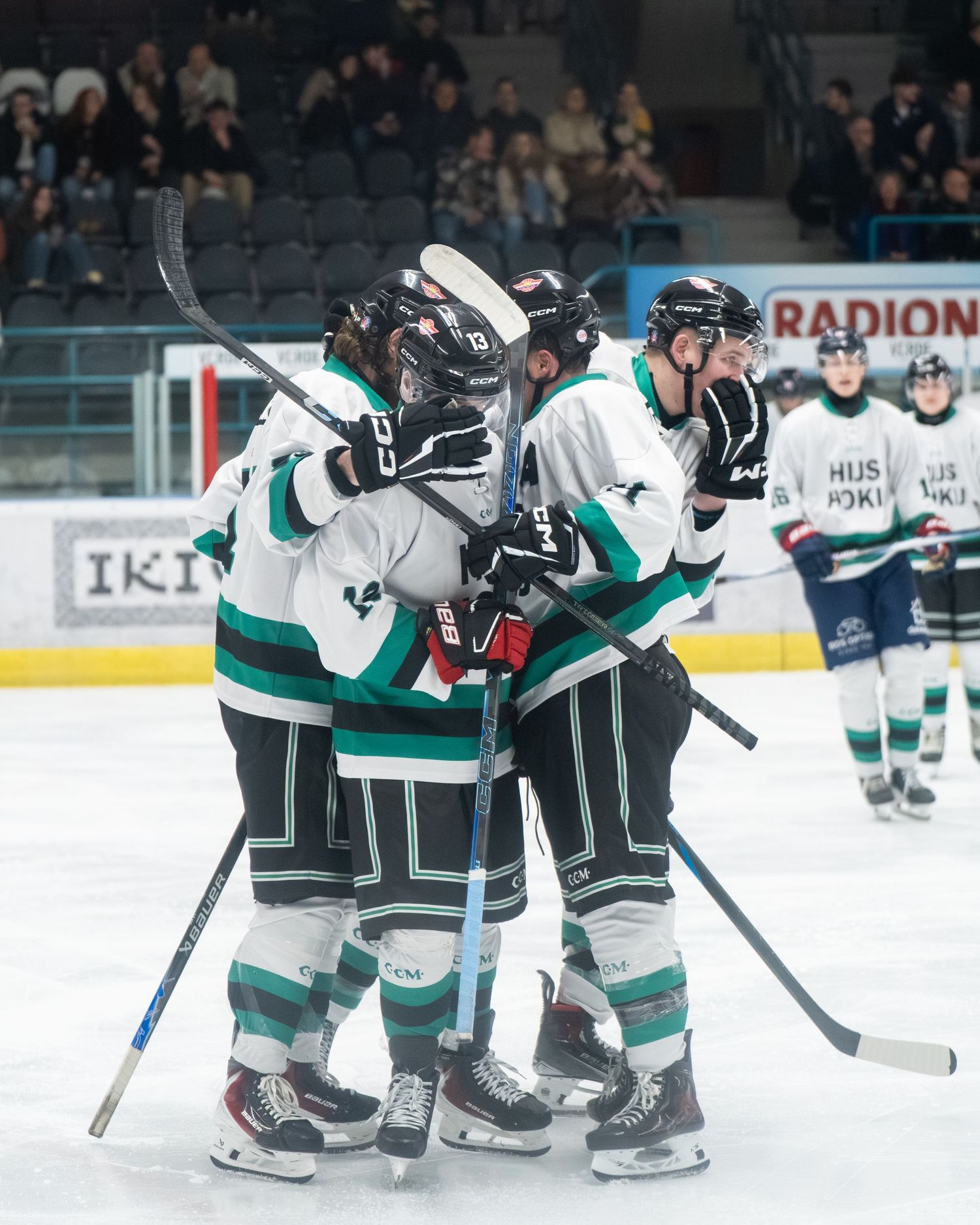 Hockey players in teal and white uniforms celebrate together on ice during a game with spectators in background.