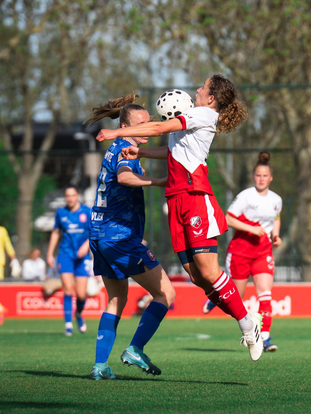 Two female soccer players battle for a header during a competitive match, one in blue and one in red and white uniforms.