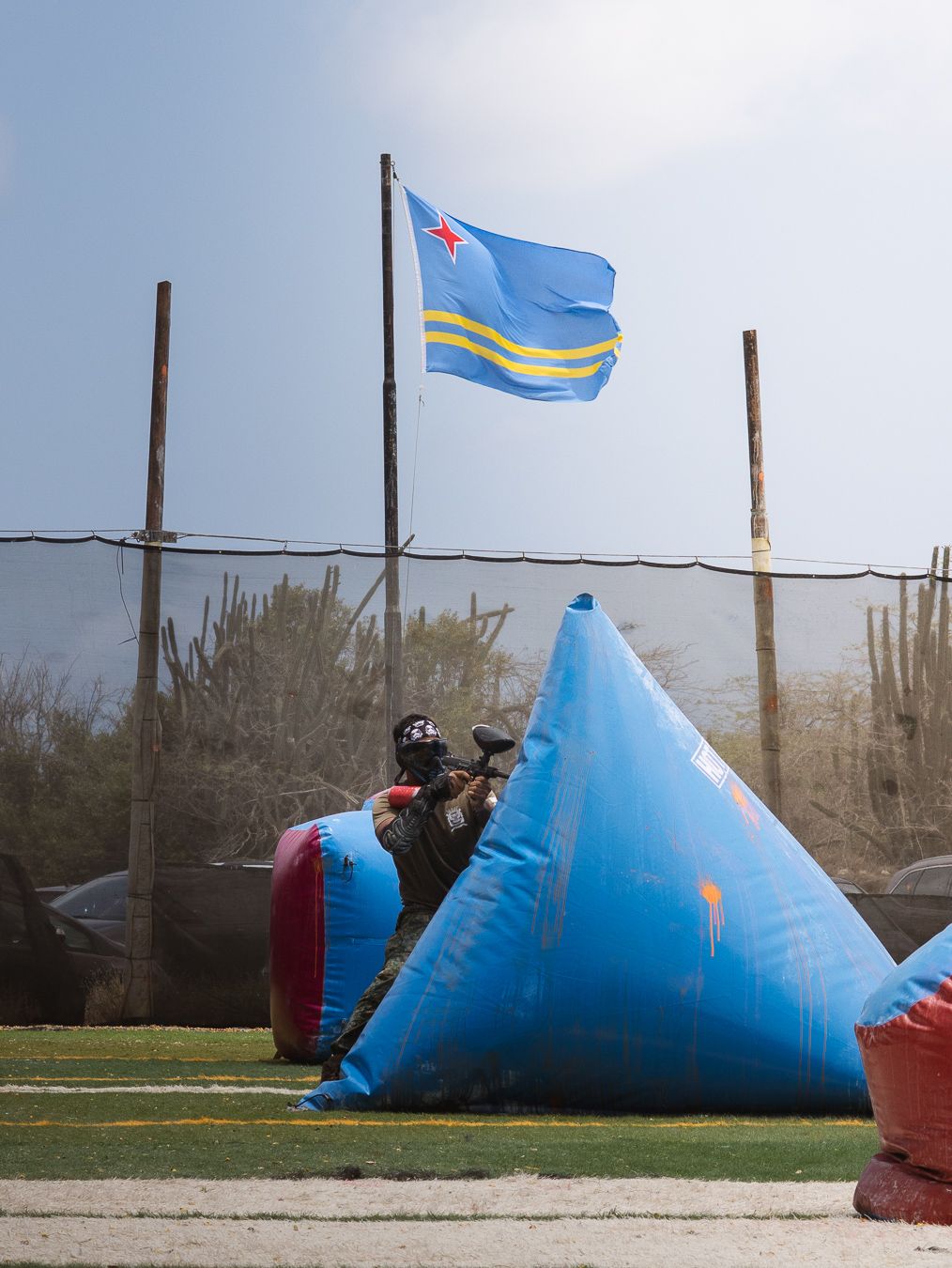 Paintball player taking cover behind inflatable bunker on field with Aruba flag flying in background.