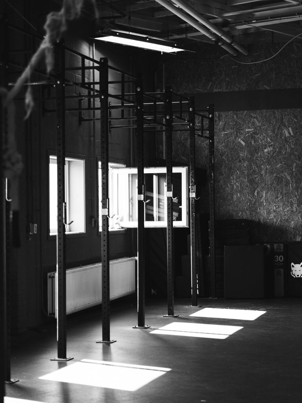 Black and white interior of a CrossFit gym showing pull-up rig stations with sunlight streaming through windows.