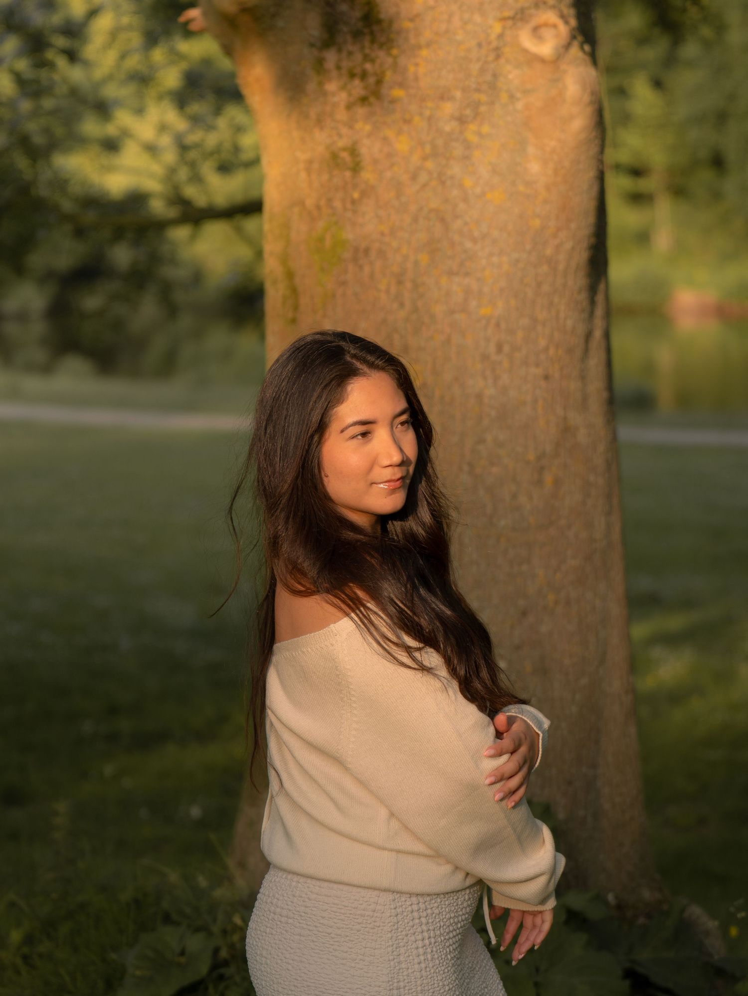 A person in a cream sweater poses against a tree trunk in warm sunset lighting at a park.