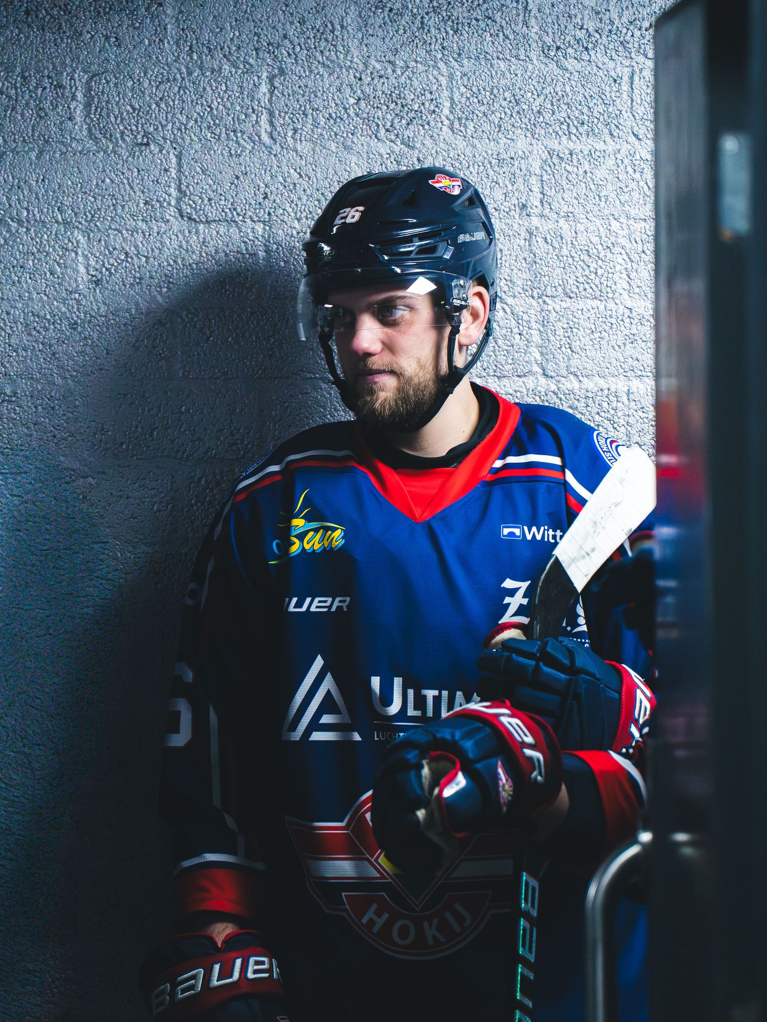 Hockey player in blue and red jersey leans against concrete wall wearing black helmet and protective gear.
