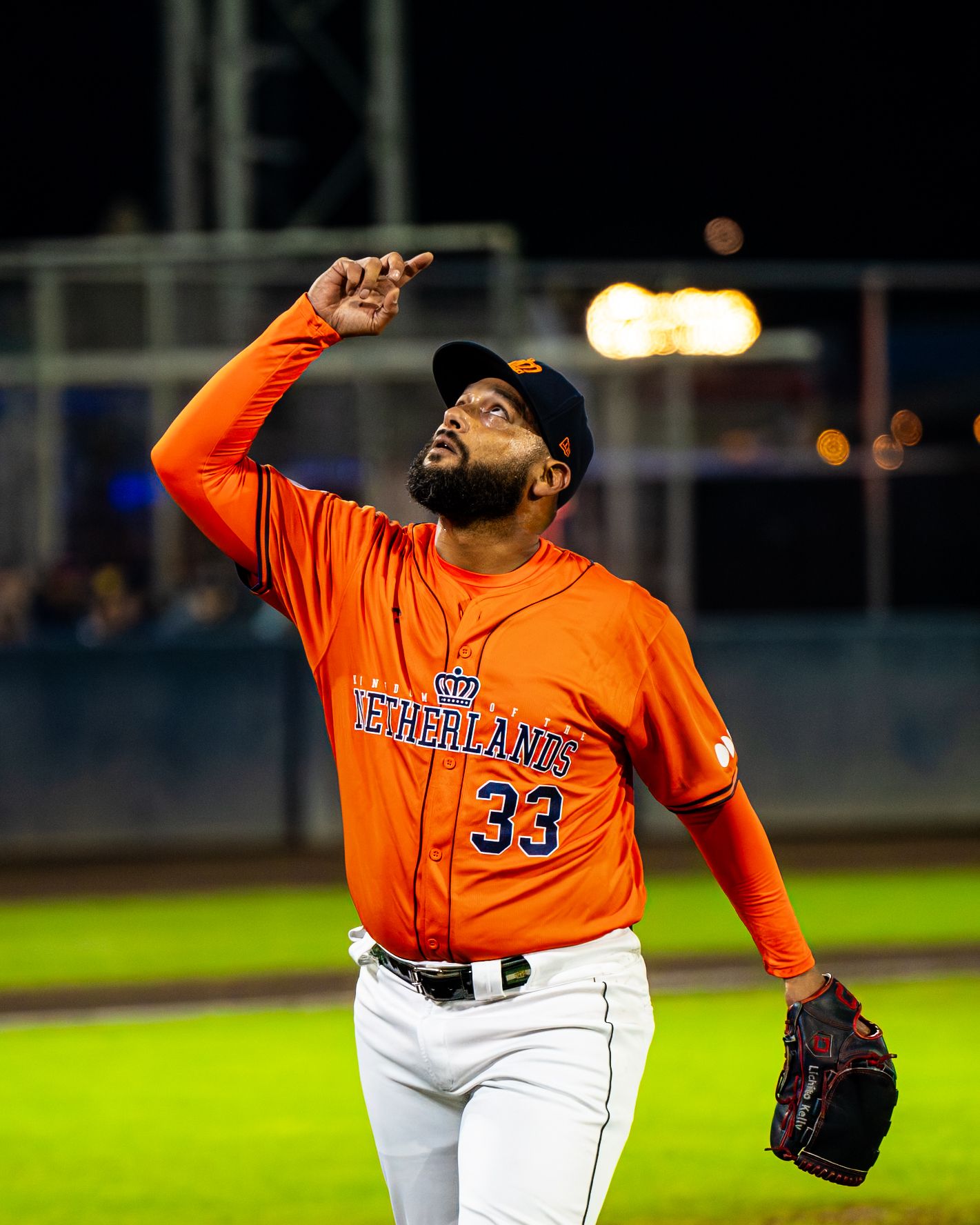 Baseball pitcher in orange jersey winds up to throw during nighttime game under stadium lights.