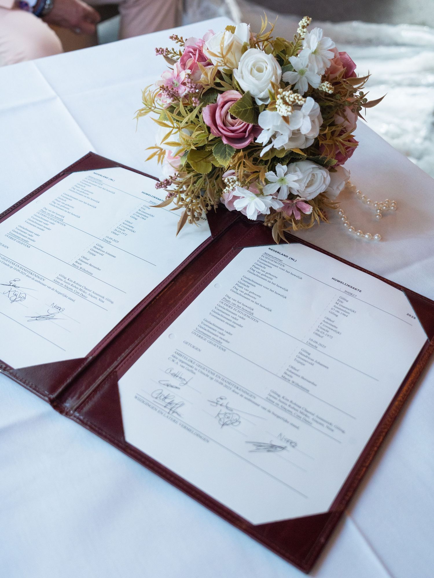 Elegant wedding menu cards with burgundy leather holders and floral arrangements in pink and white on a white tablecloth.