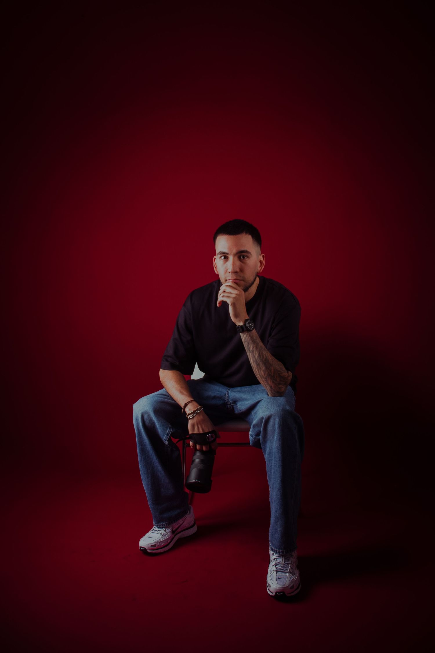 A figure in dark clothing sits crouched in a moody studio setting with dramatic red lighting and shadows.