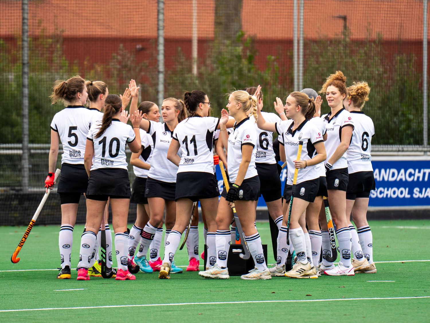 Field hockey team in white uniforms huddles together on artificial turf field before a match.