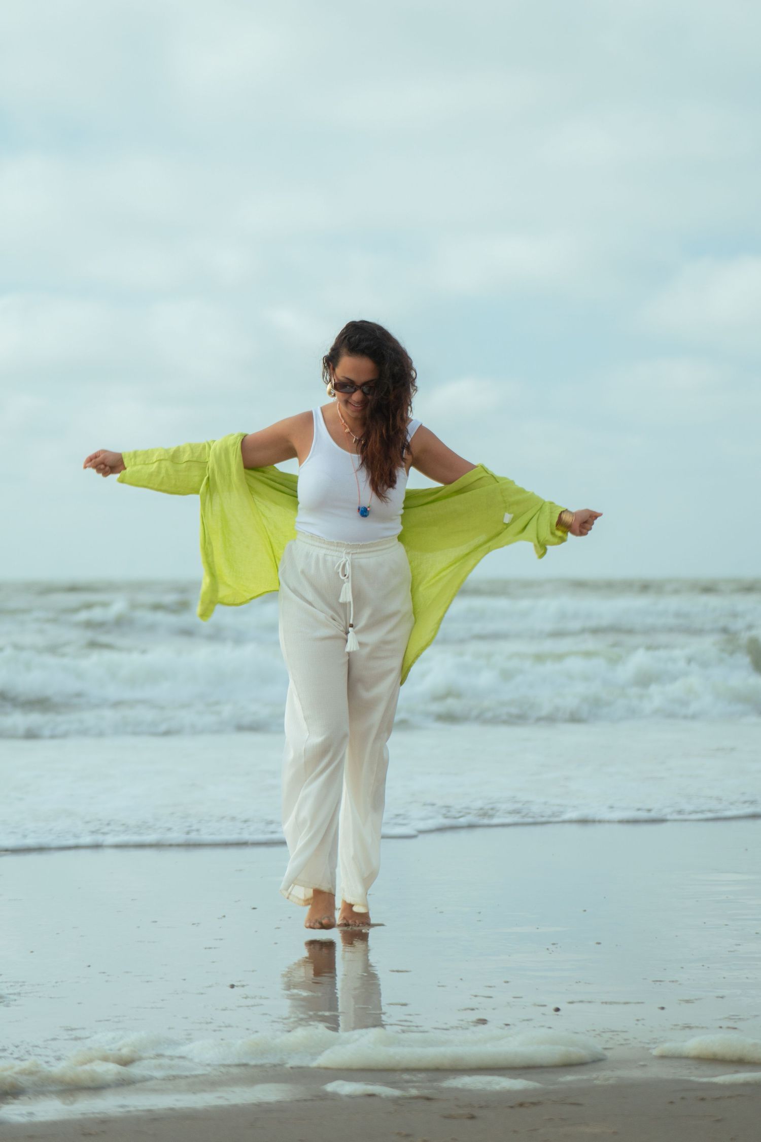 Woman in white pants and neon yellow cardigan dancing joyfully on a cloudy beach.