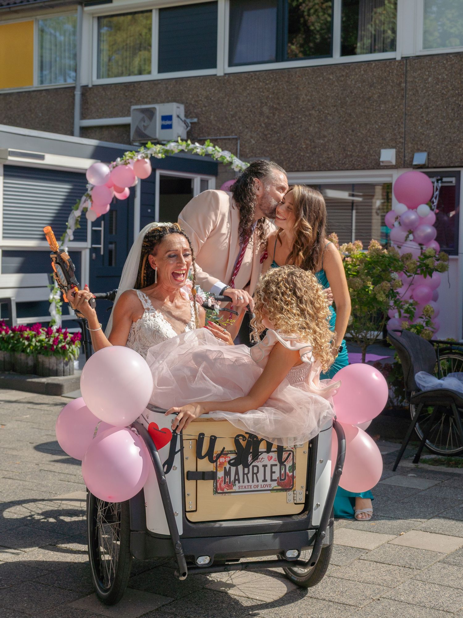 Three photos showing people celebrating with pink balloons and a cargo bike on a sunny day outside a building.