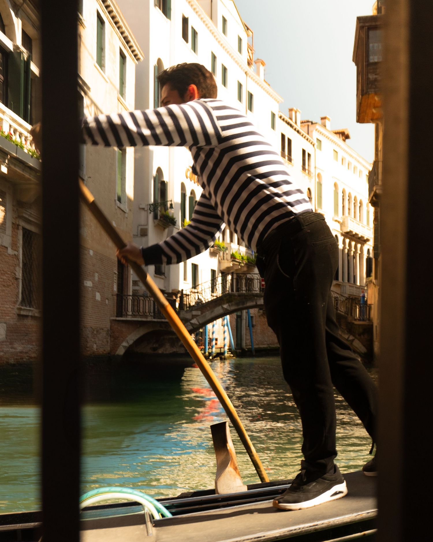 A gondolier in a striped shirt skillfully navigates a wooden oar through a sunlit Venice canal.