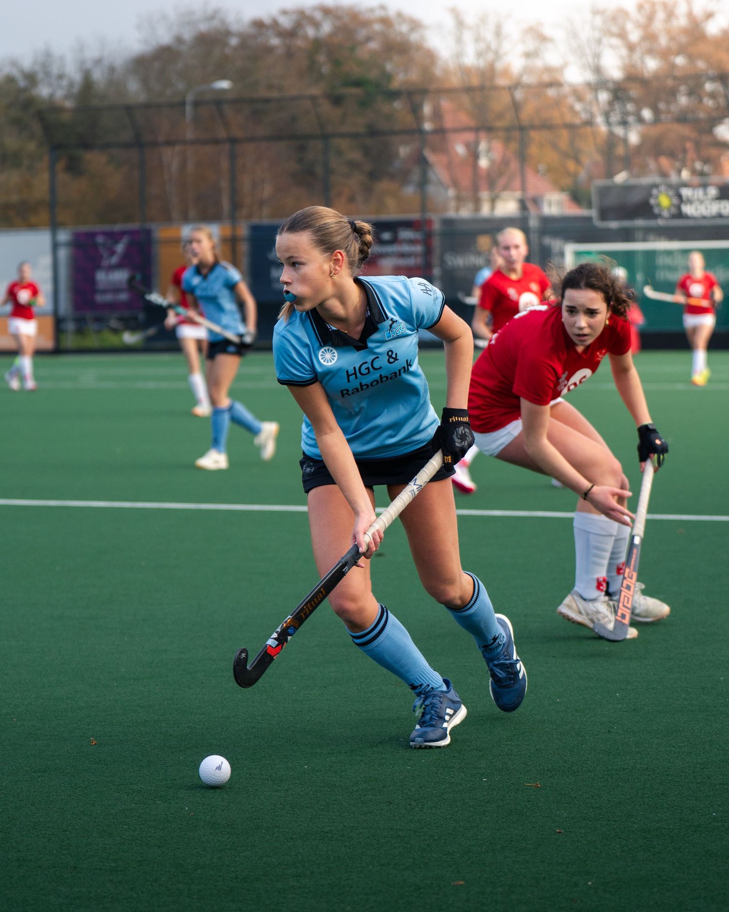 Field hockey players in blue and red uniforms compete for the ball during an intense match on artificial turf.