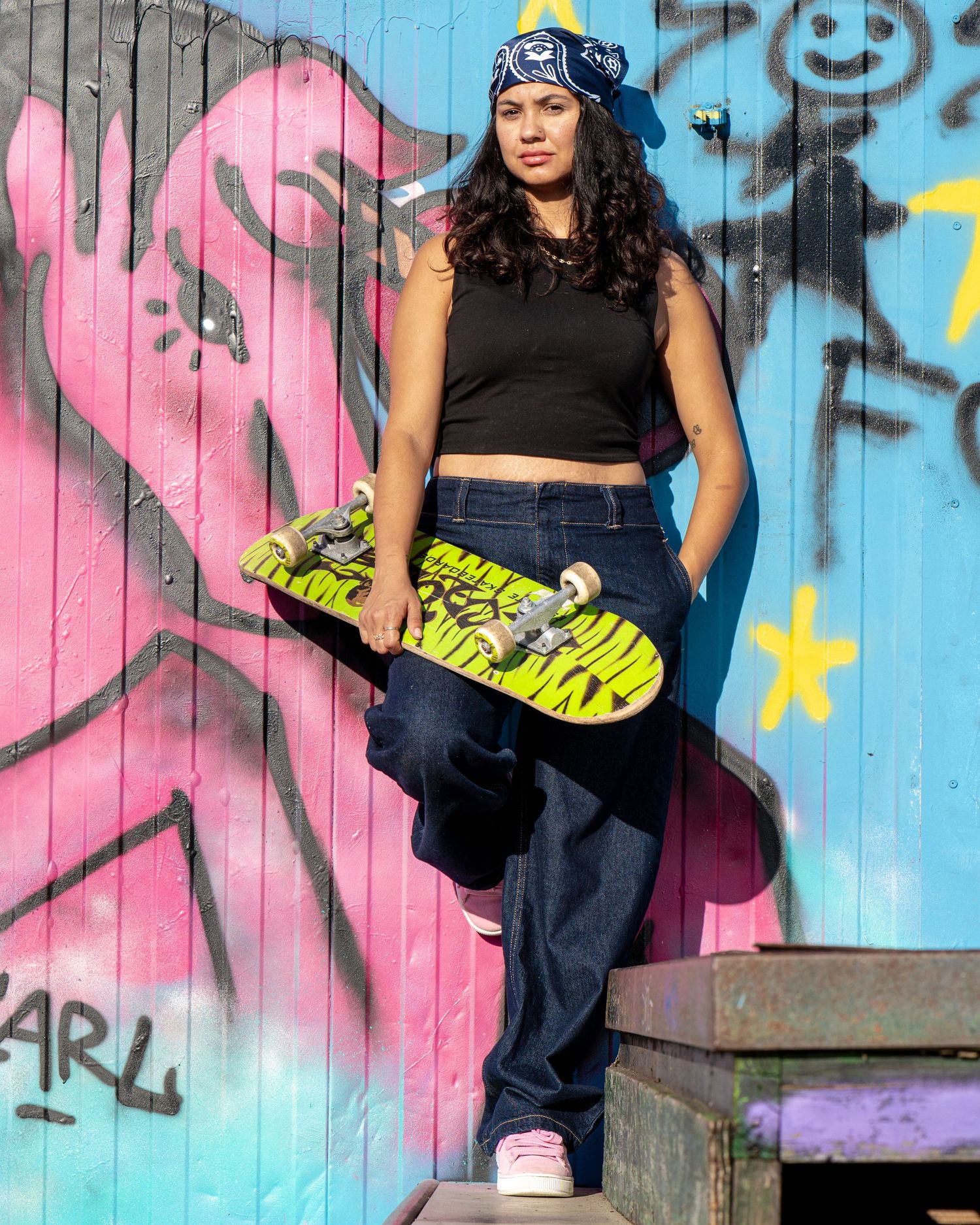 Skateboarder in blue beanie and baggy jeans poses with skateboard against colorful graffiti wall in urban setting.