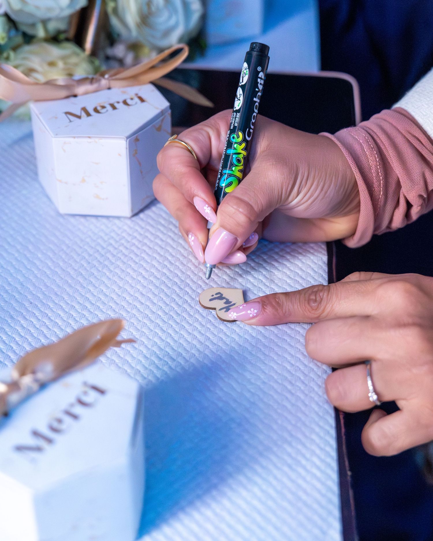 Hand writing on note card next to white 'Merci' favor box at wedding reception.
