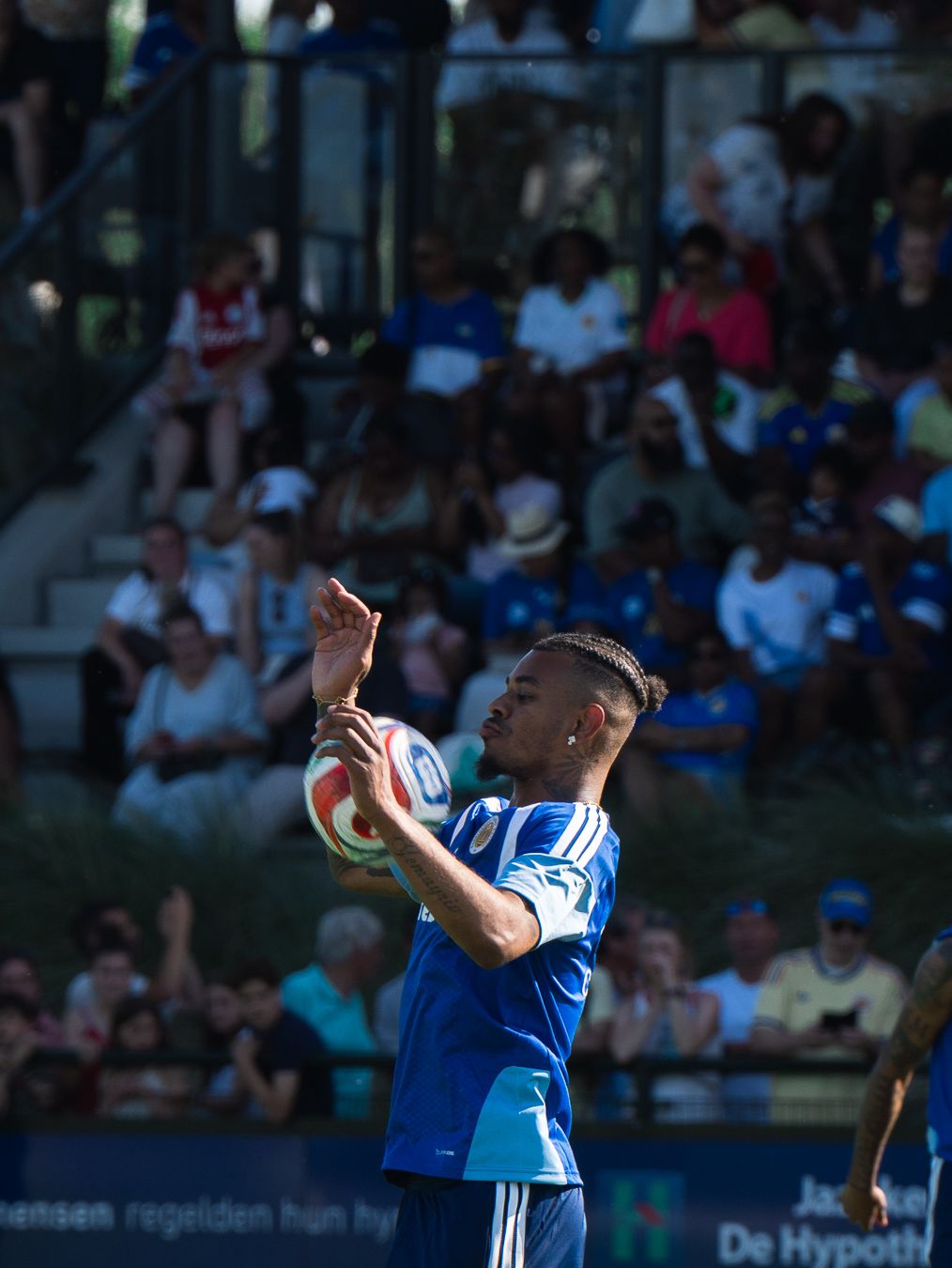 Soccer player in blue Curaçao kit holds ball during a sunny outdoor match with spectators in background.