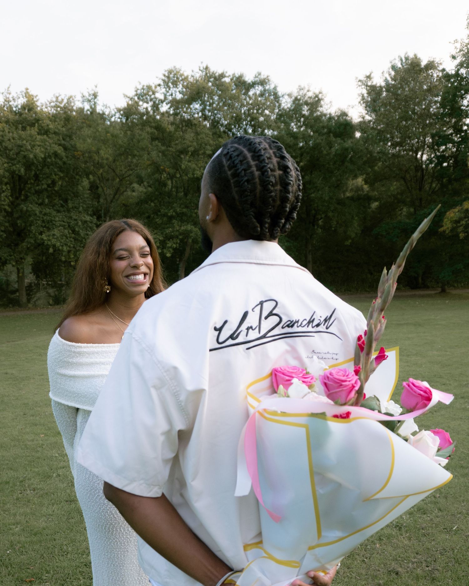 A couple in white outfits embrace in a romantic outdoor photoshoot with pink floral accents.