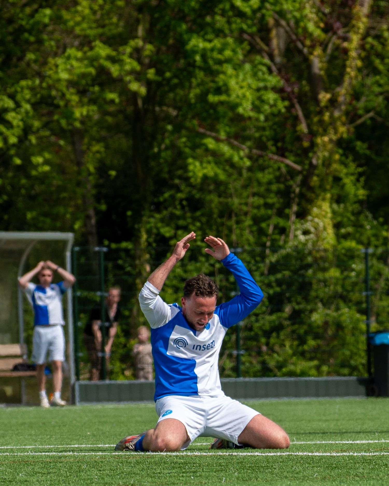 Soccer player falls dramatically onto artificial turf field during outdoor match with trees in background.