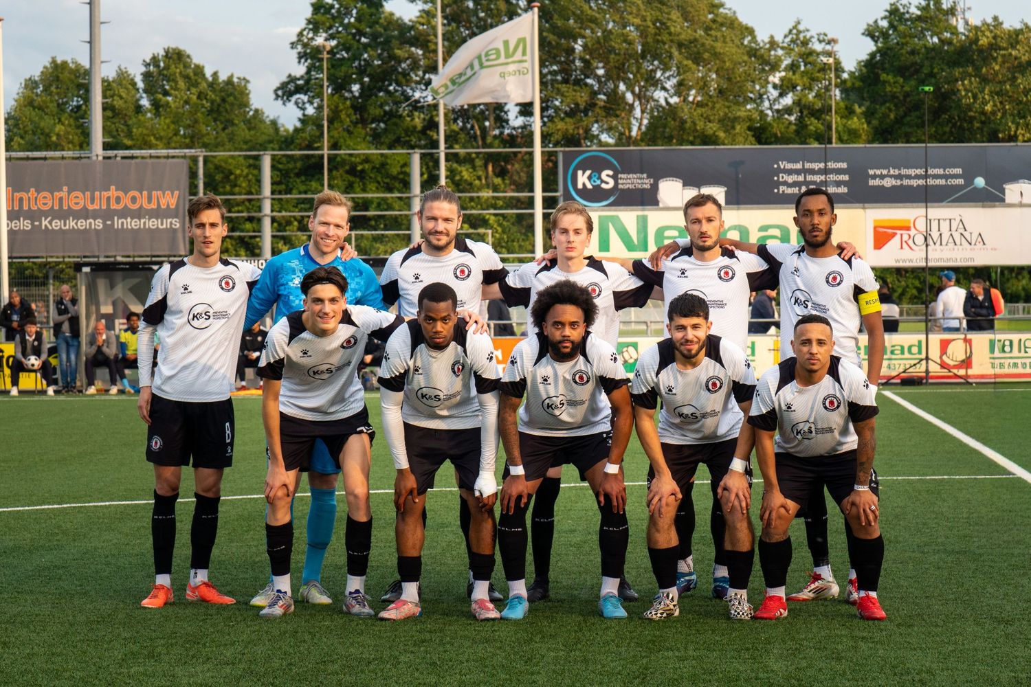 A soccer team poses for a group photo on a green field during an evening match in a Dutch amateur football league.