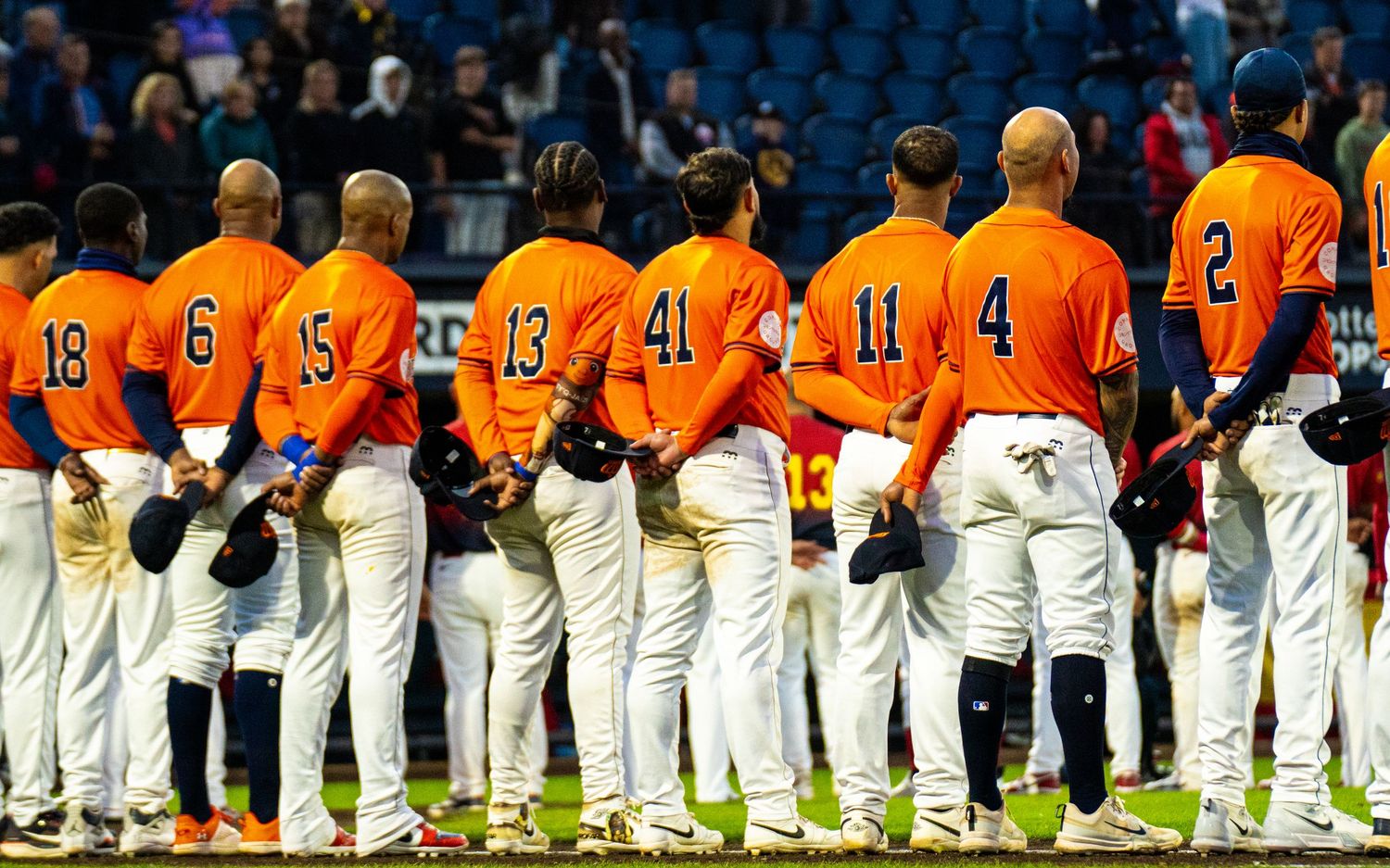 Baseball team in orange uniforms lined up on field during pre-game ceremony, players wearing numbers 18, 6, 15, 13, 41, 11, 4, 2.