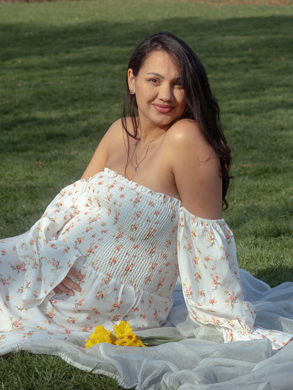 Woman in white floral off-shoulder dress sitting on grass with yellow flowers, smiling outdoors.