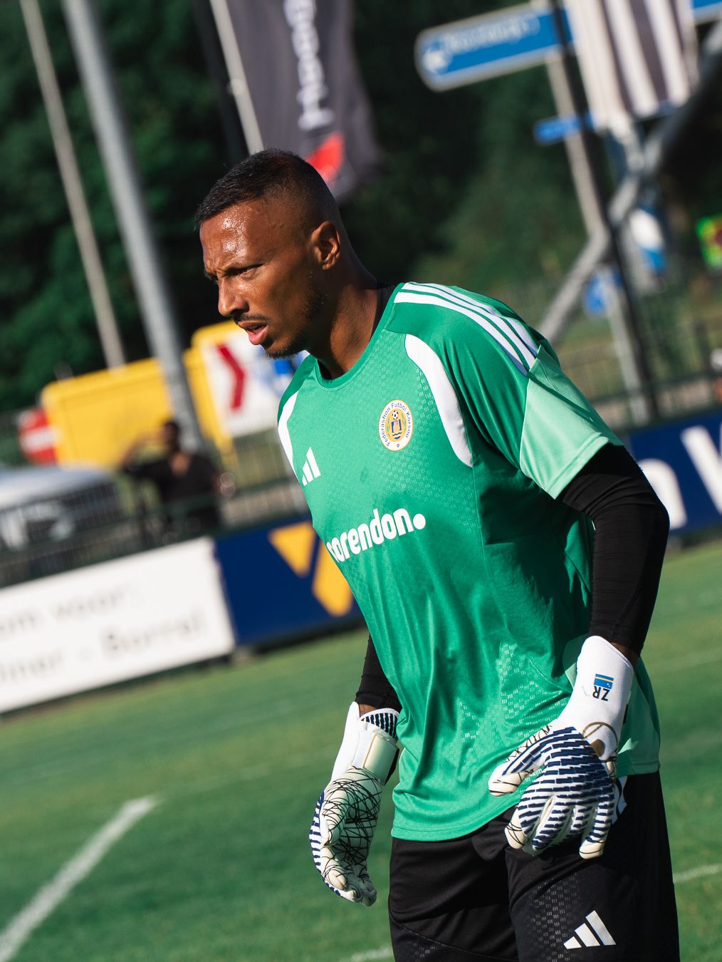 A soccer goalkeeper in a green Adidas jersey and gloves stands on a training pitch during a football session.