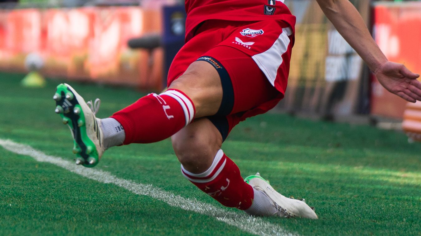 Soccer player in red and white uniform kicking a ball on green grass field wearing white cleats.