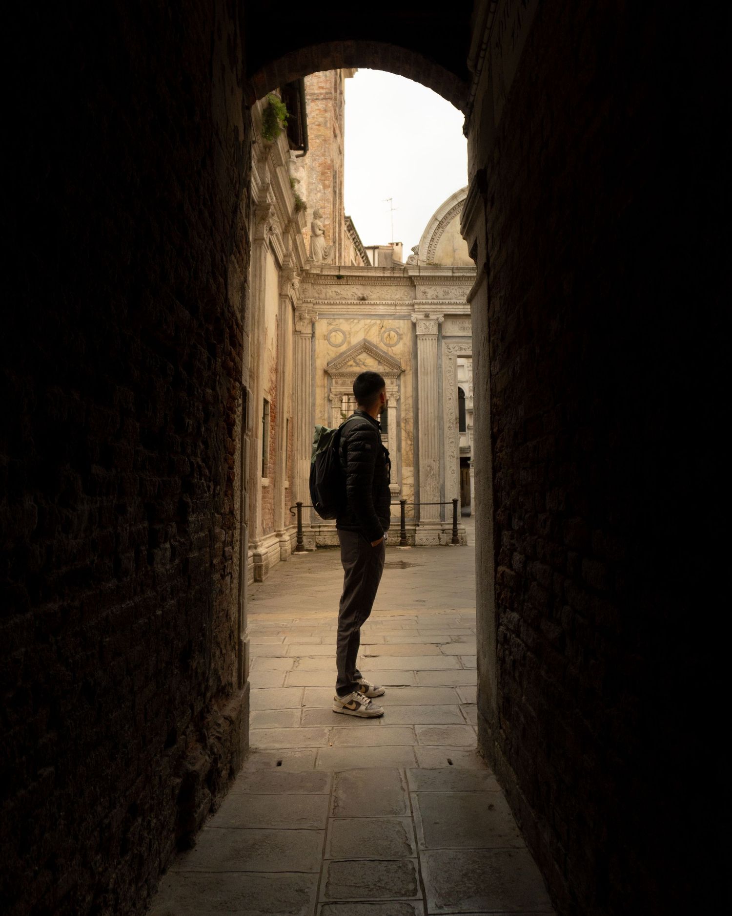 Man with backpack standing in a dark stone archway, gazing at a sunlit historic European courtyard.