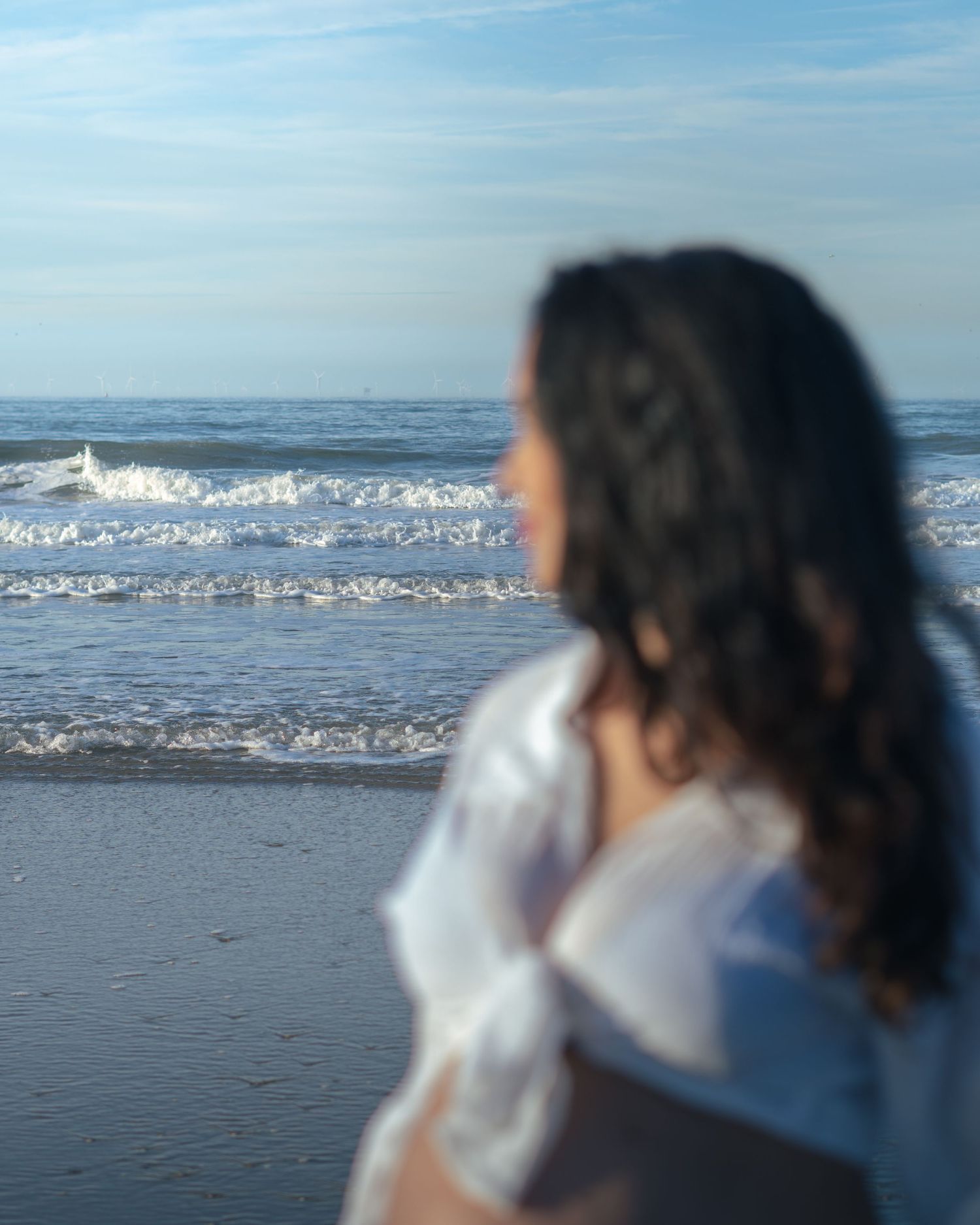 A woman with curly dark hair in a white shirt gazes at ocean waves on a sandy beach under a blue sky.