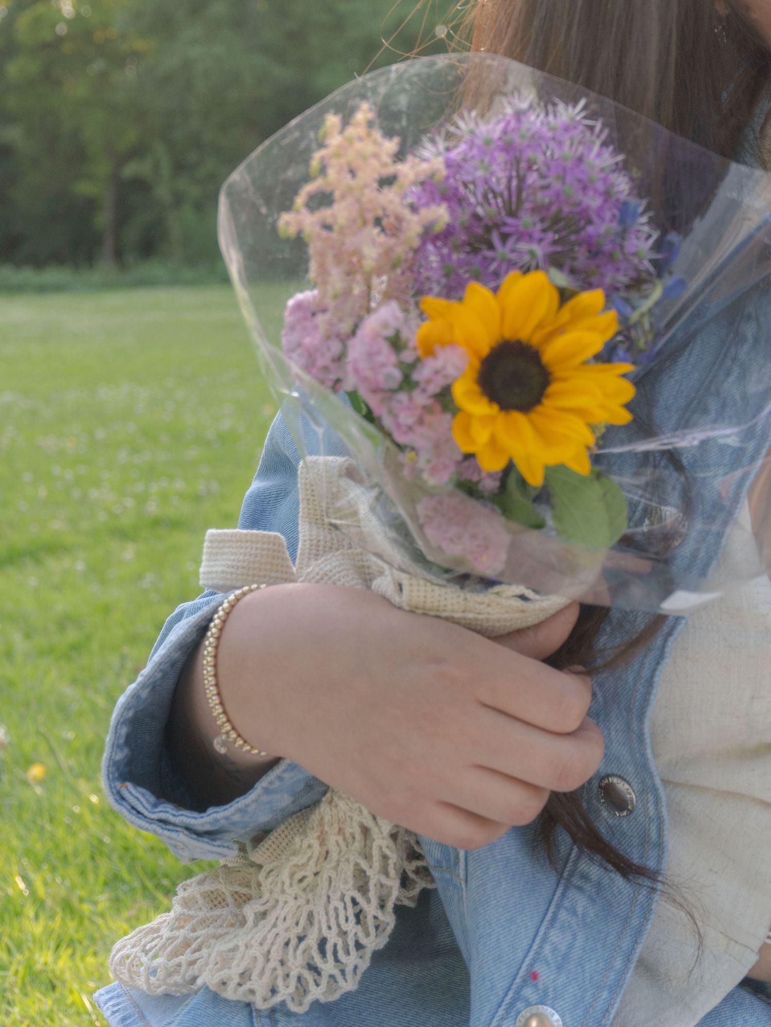 A bouquet with a bright sunflower, purple allium, and peach flowers wrapped in cellophane is held against a grassy background.