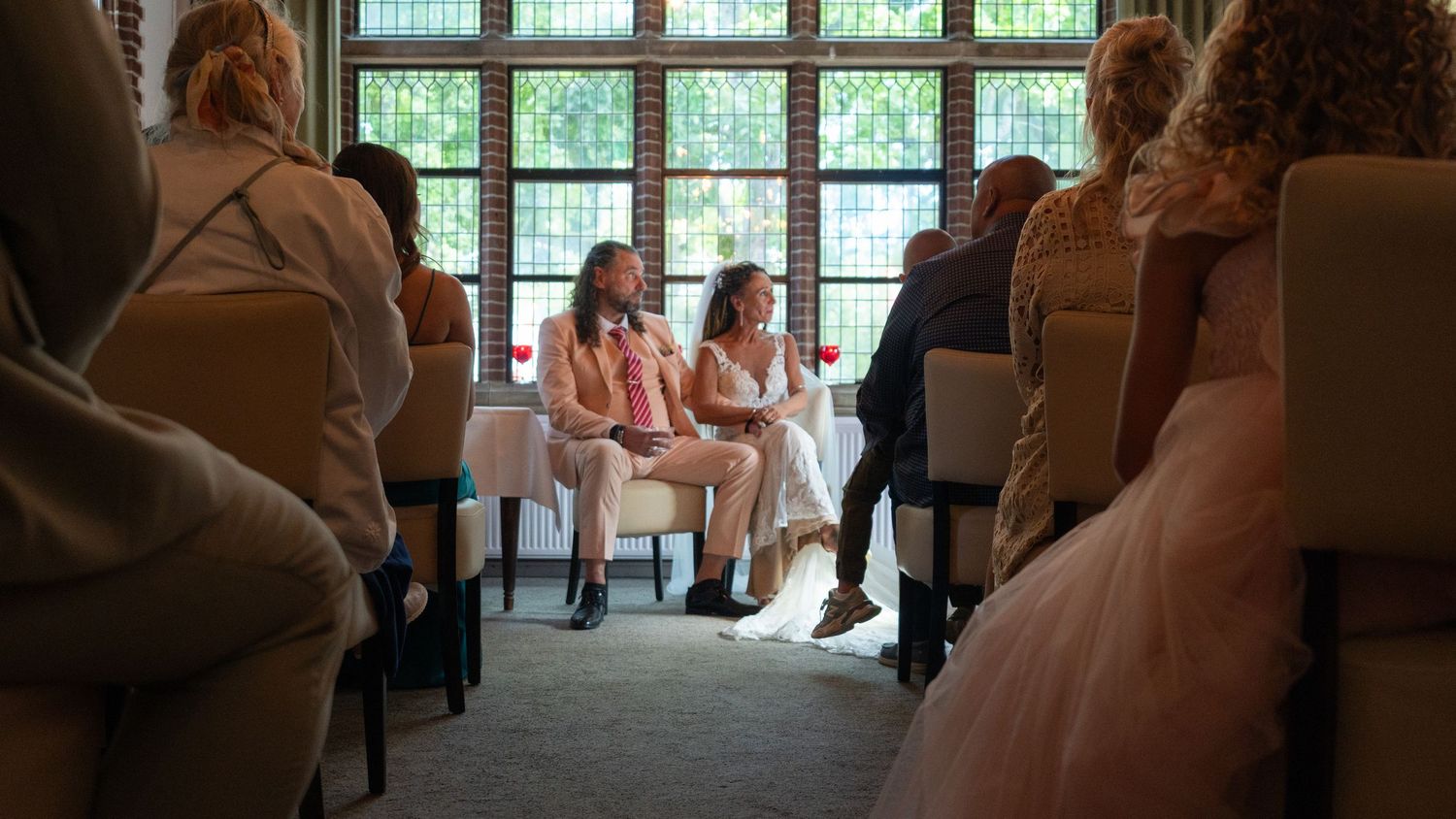 Two people sit together in conversation during a formal event with guests seated in elegant chairs.