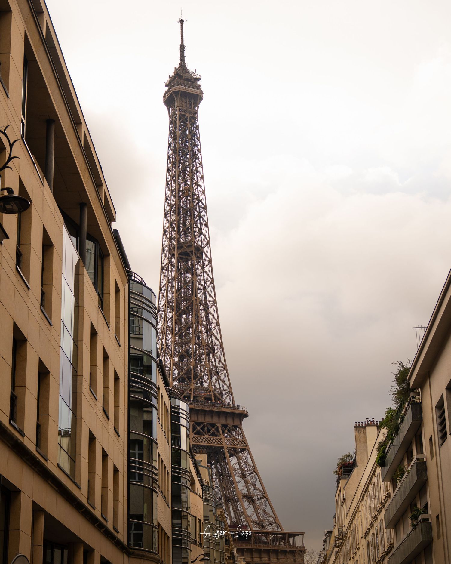 Eiffel Tower rising above Parisian streets between classic Haussmann buildings under dramatic cloudy skies.