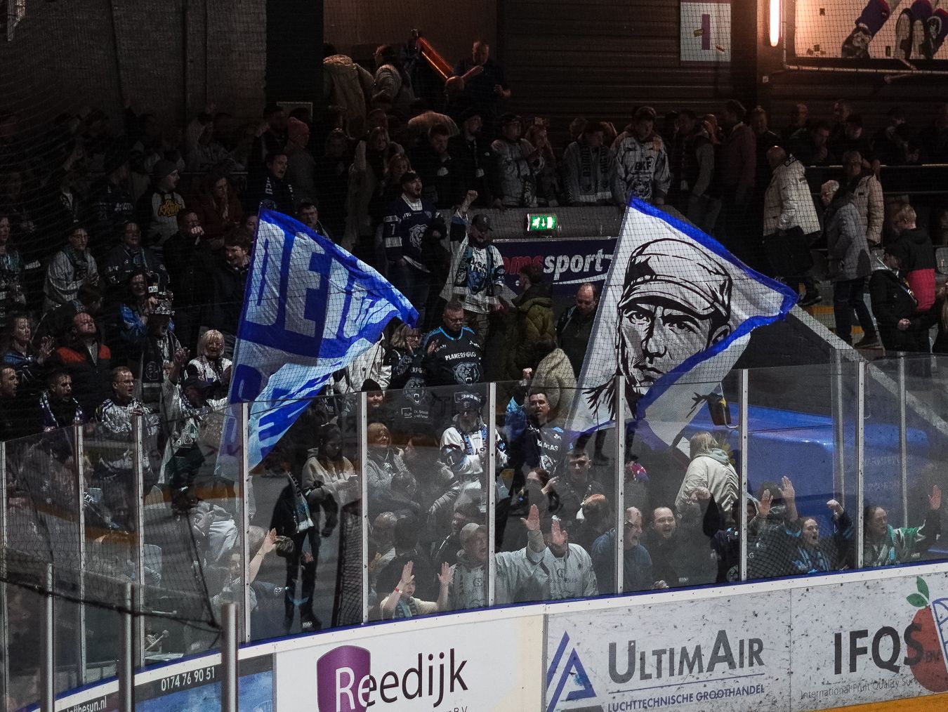 Ice hockey fans wave team flags behind the glass at an indoor arena with sponsor boards visible.