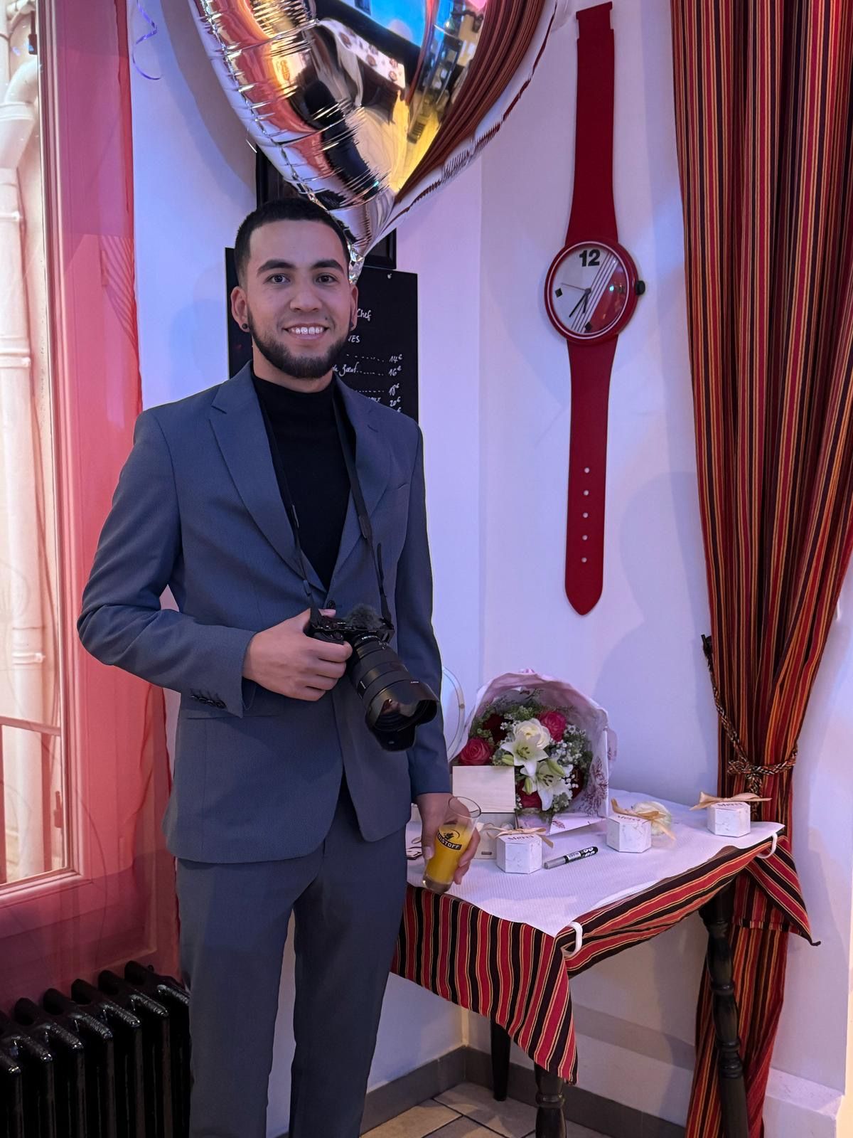 A person in a gray suit and black turtleneck stands smiling in an elegant room with curtains and a wall clock.