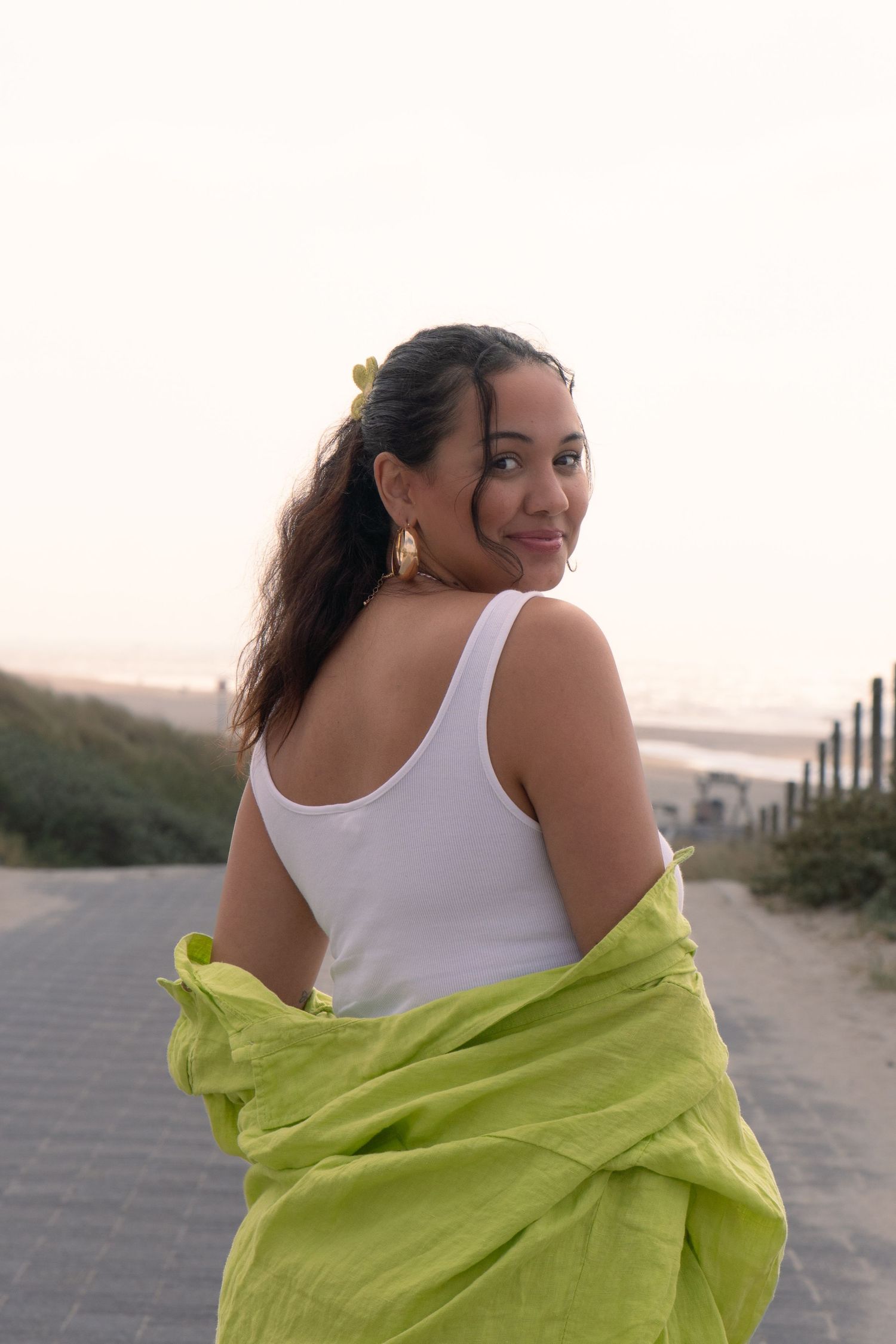Woman in white tank top and lime green wrap poses on beach boardwalk at sunset.