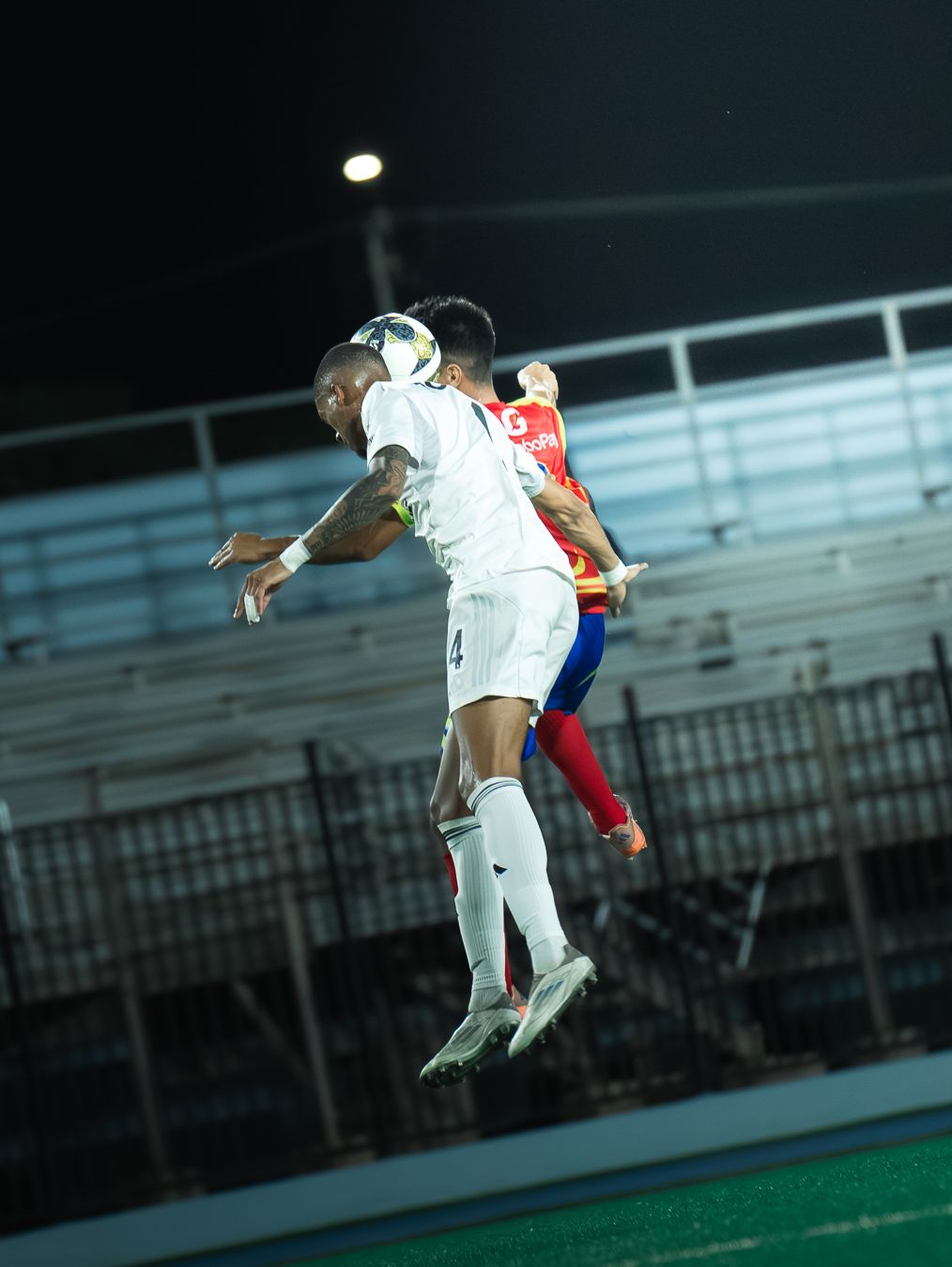 Two soccer players jump to head the ball during a night match, one in white and one in red uniforms.